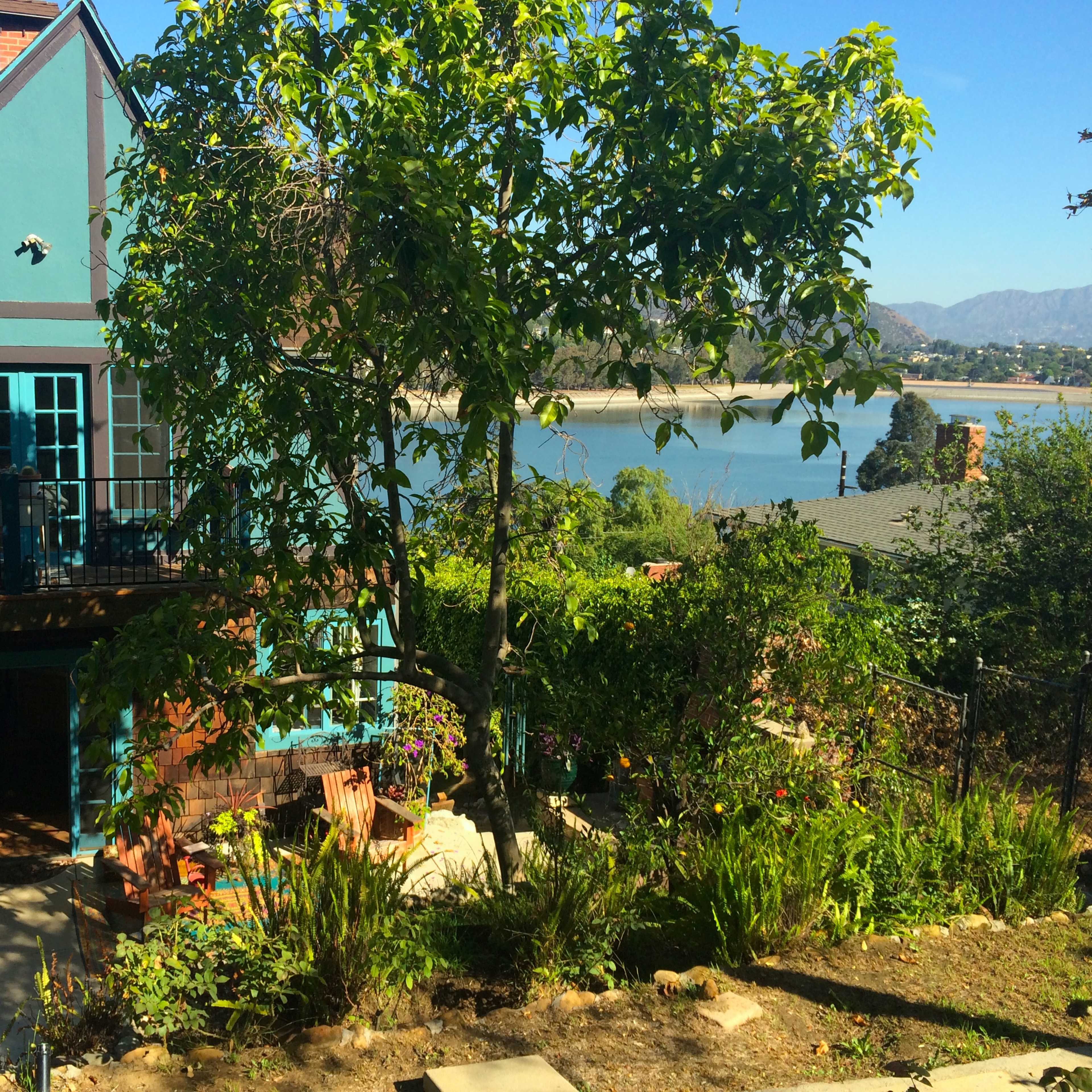 A blue and brown house with a balcony overlooks a body of water surrounded by mountains and greenery.
