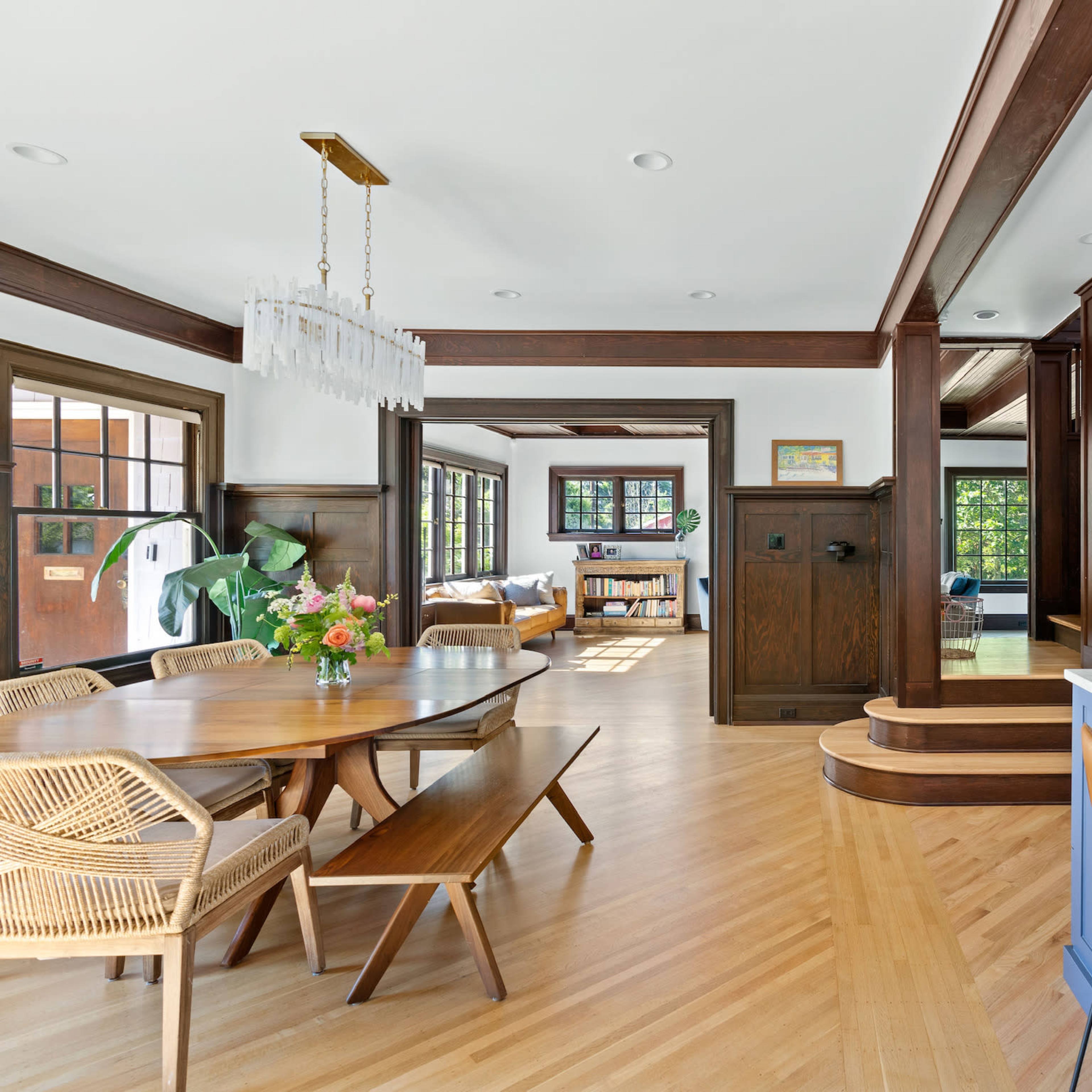 A modern dining area with a wooden table, rattan chairs, and a kitchen with blue cabinetry and a marble countertop, alongside a sunlit living space.