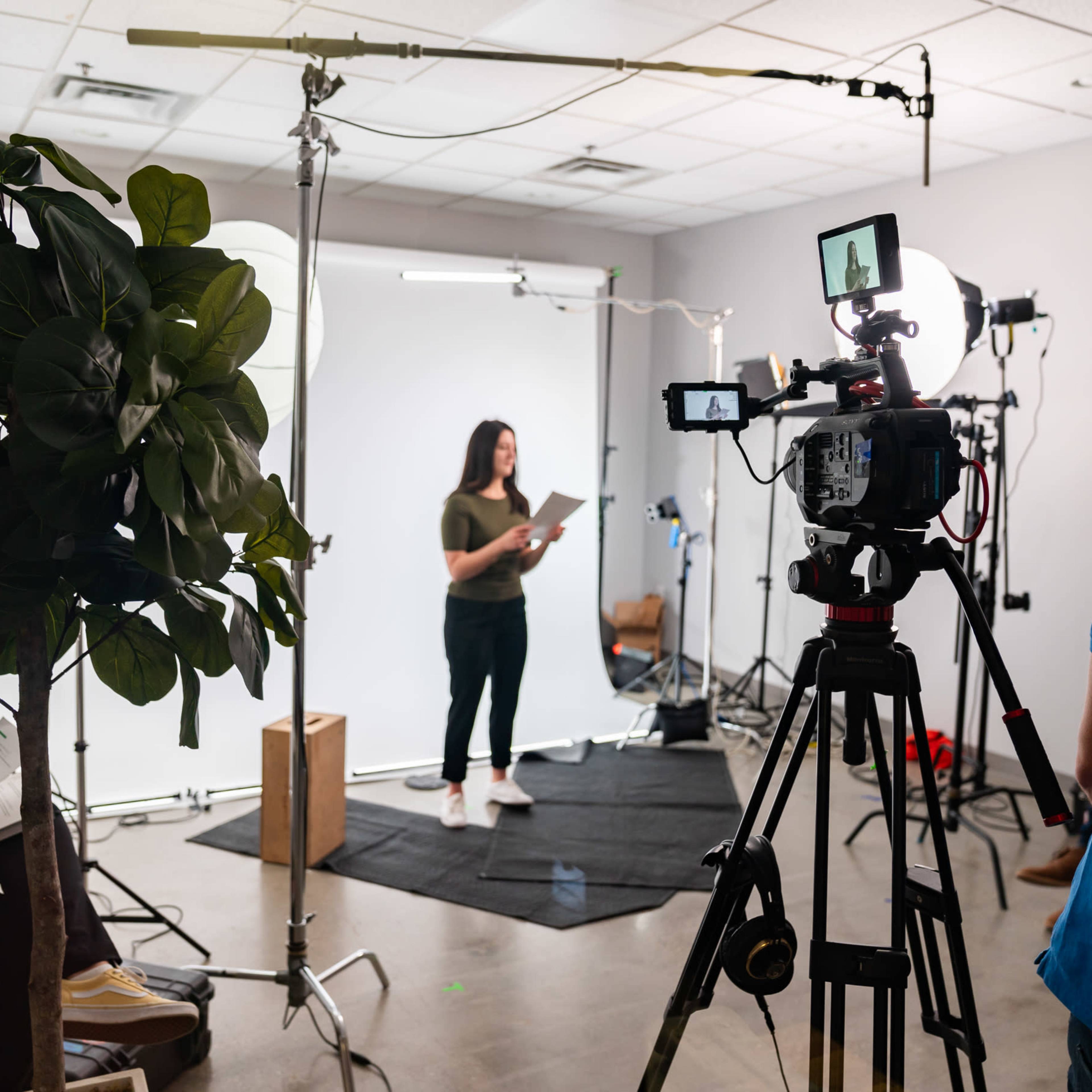 A woman is standing on a black mat in front of a white backdrop while holding a script, surrounded by video equipment and studio lights.