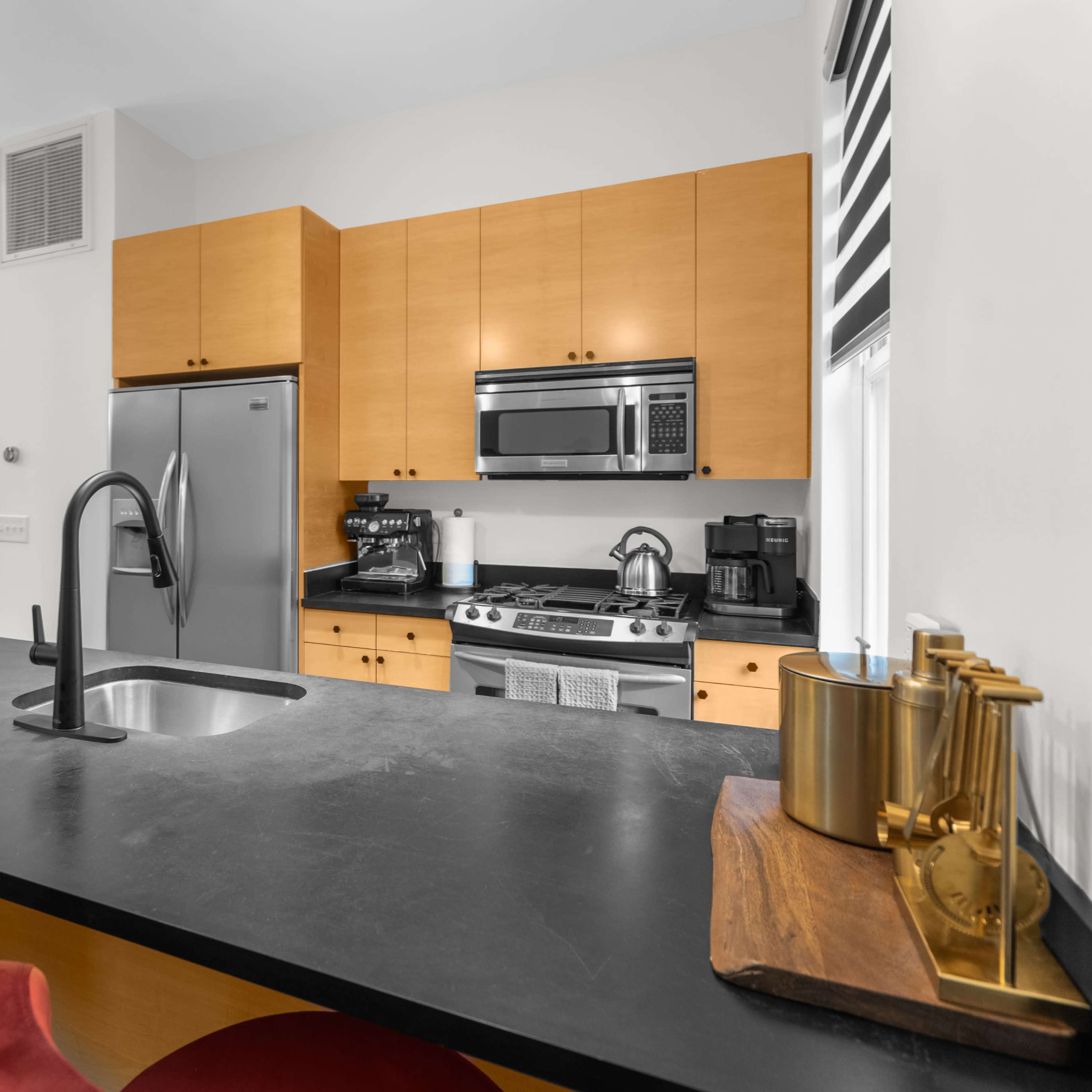 A modern kitchen features wooden cabinets, stainless steel appliances, and a black countertop with a sink and red bar stools.