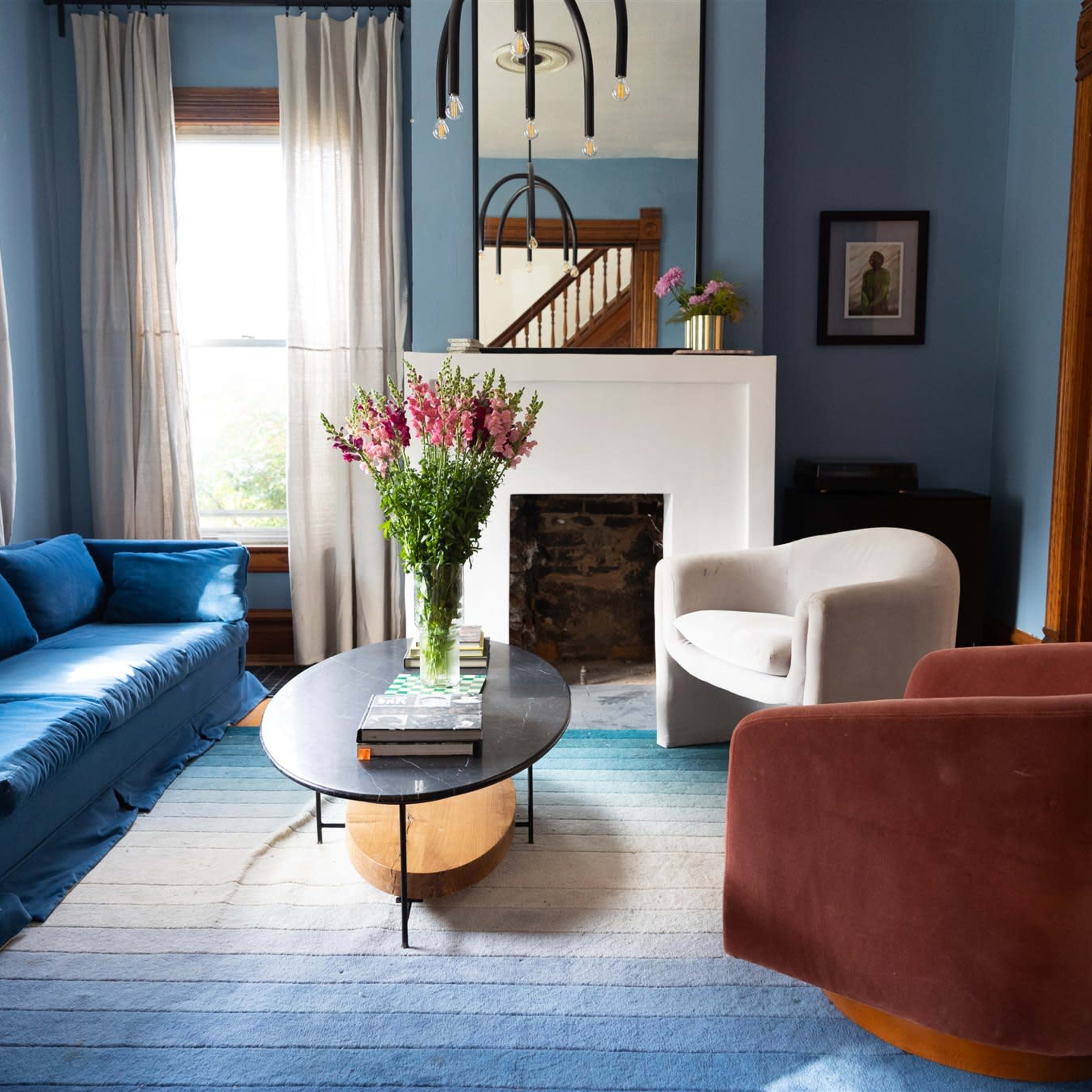 A living room with a blue couch, a white chair, a circular coffee table, and flowers in a vase on top of the table, with large windows providing natural light.