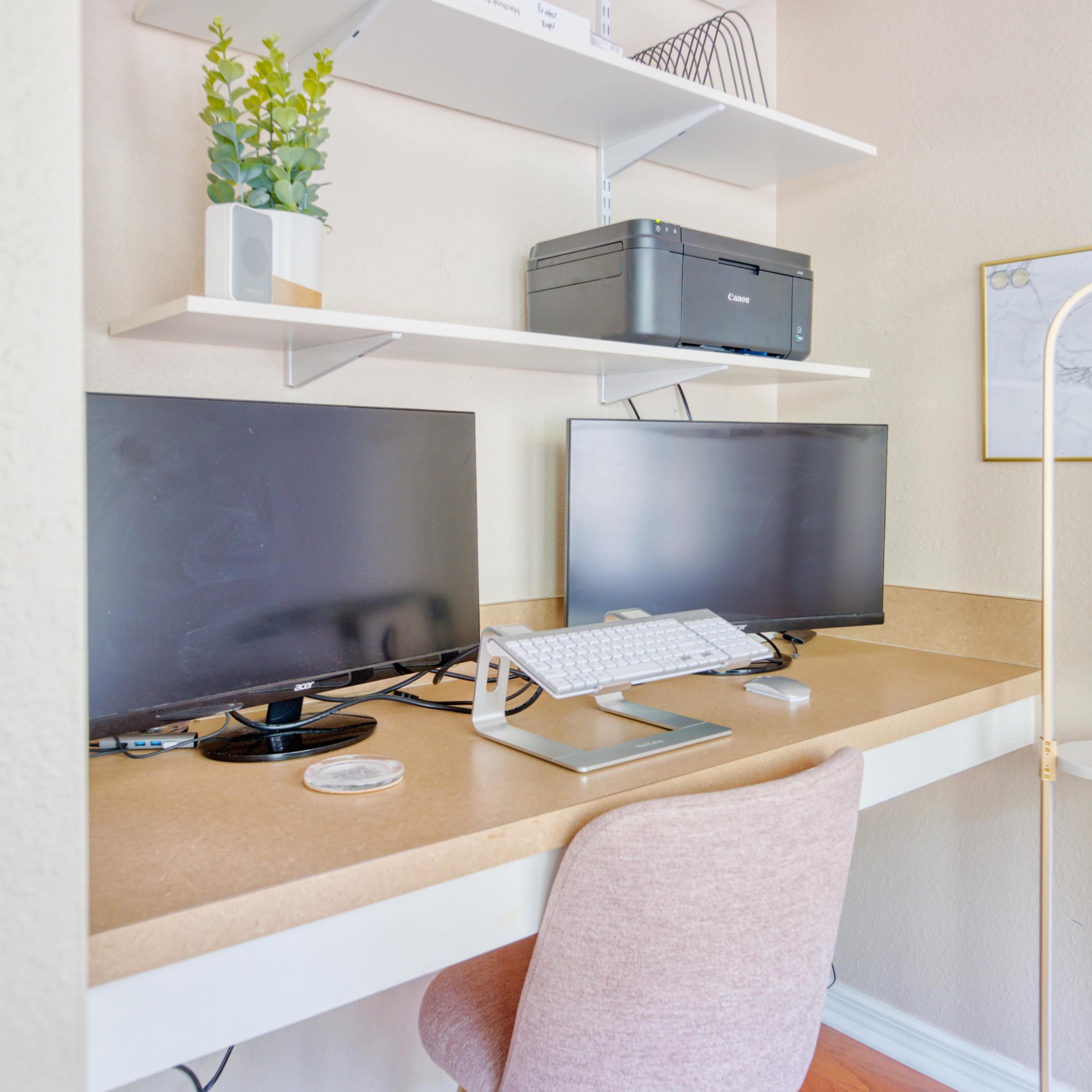 A workspace featuring two computer monitors, a keyboard on a stand, and a pink chair against a light-colored wall with shelves above holding decorative plants and a printer.