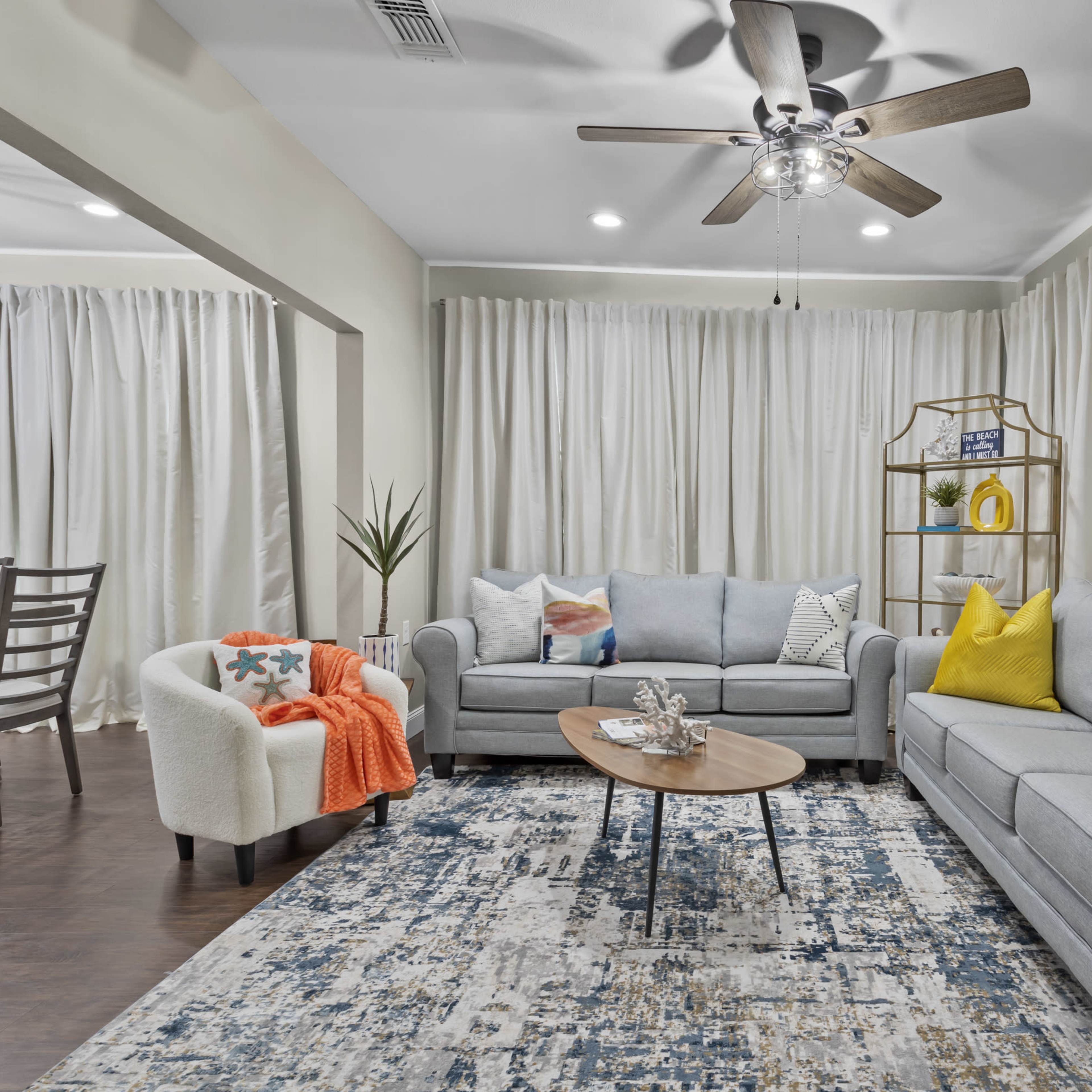 A modern living room features a gray sectional sofa, a round wooden coffee table, and an area rug, with light-colored curtains and a dining area visible in the background.