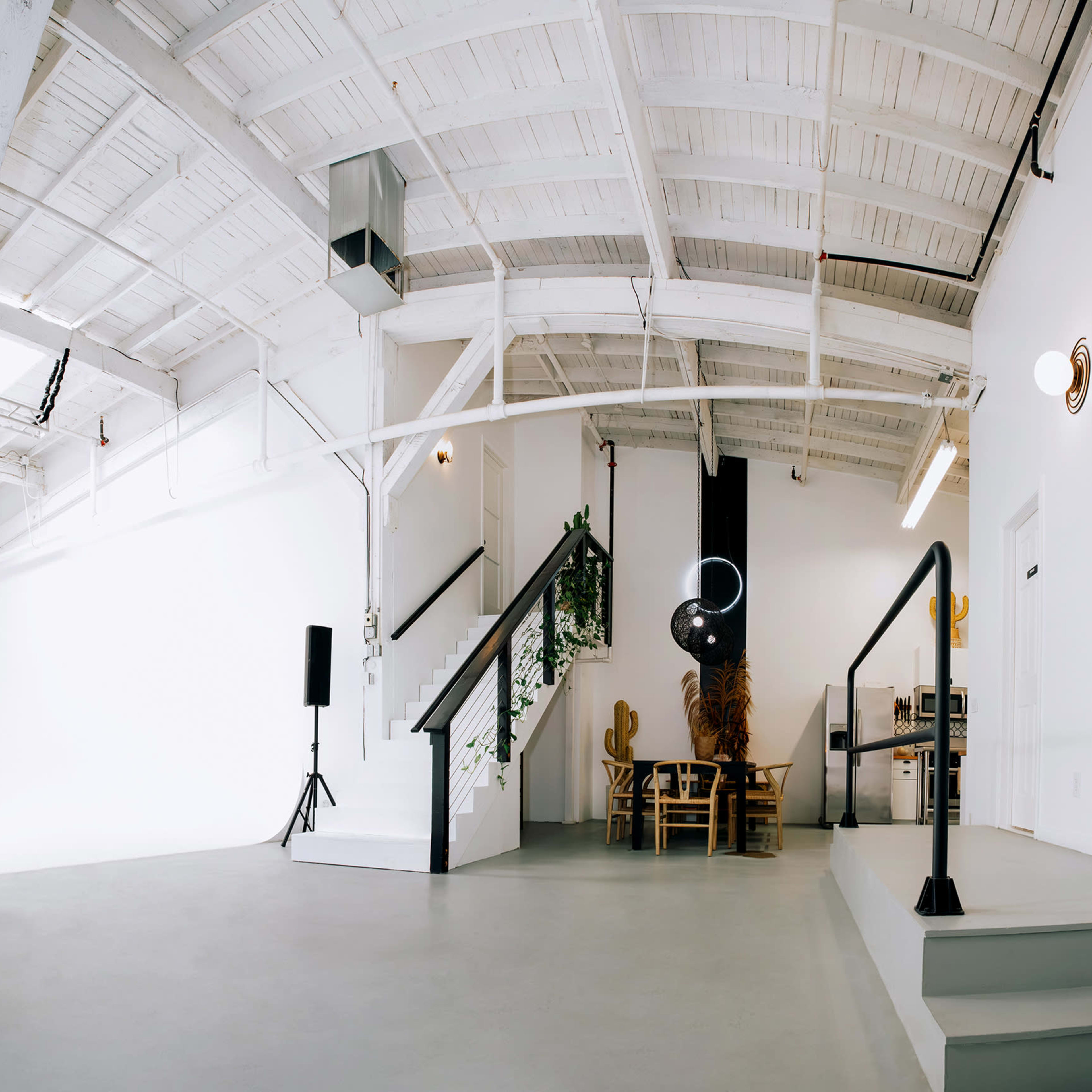 A spacious, well-lit studio with a high ceiling, white walls, and minimalistic decor, featuring speakers, lighting equipment, and a staircase leading to a small seated area.
