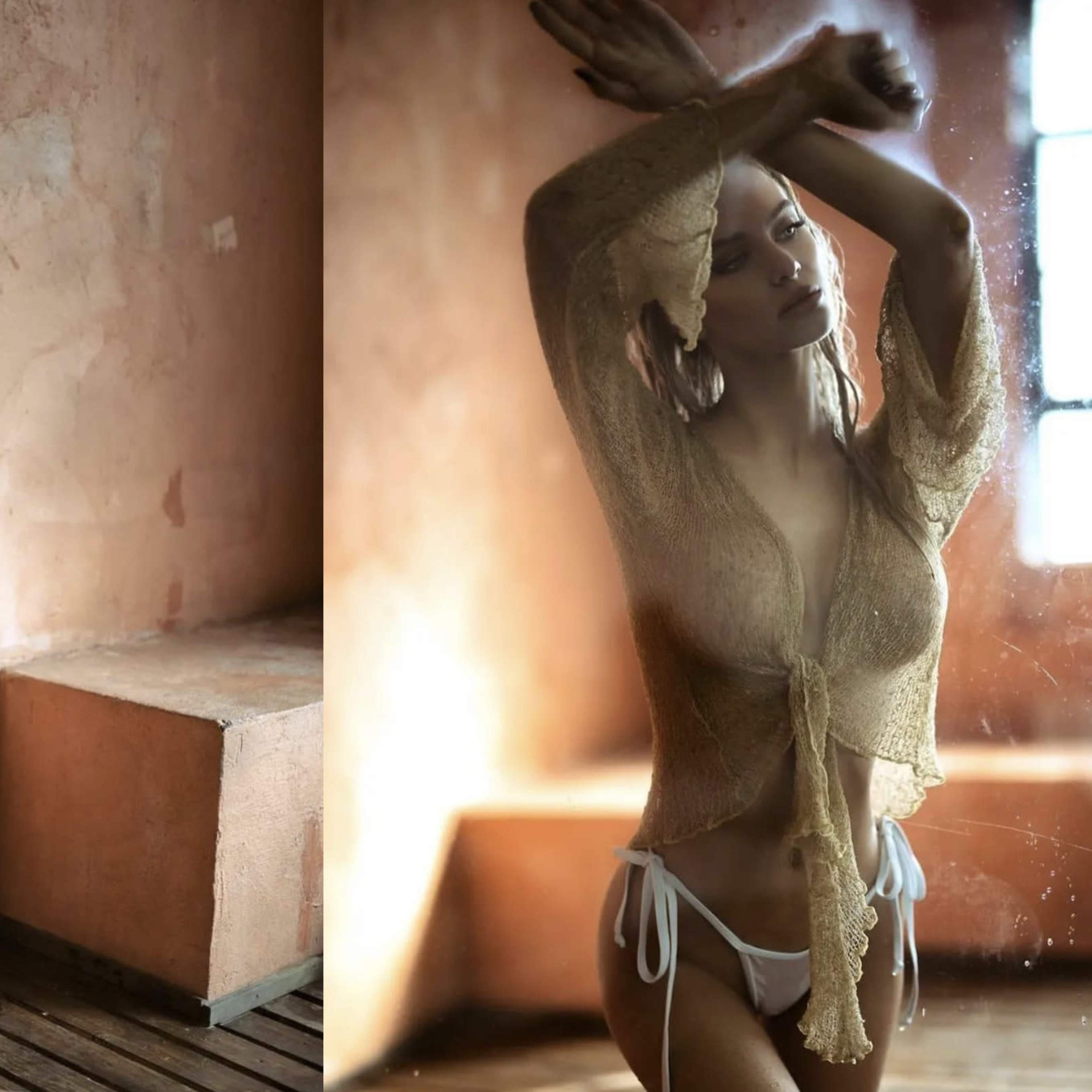 A person poses in a softly lit room with textured walls, wearing a sheer top and minimal swimwear.
