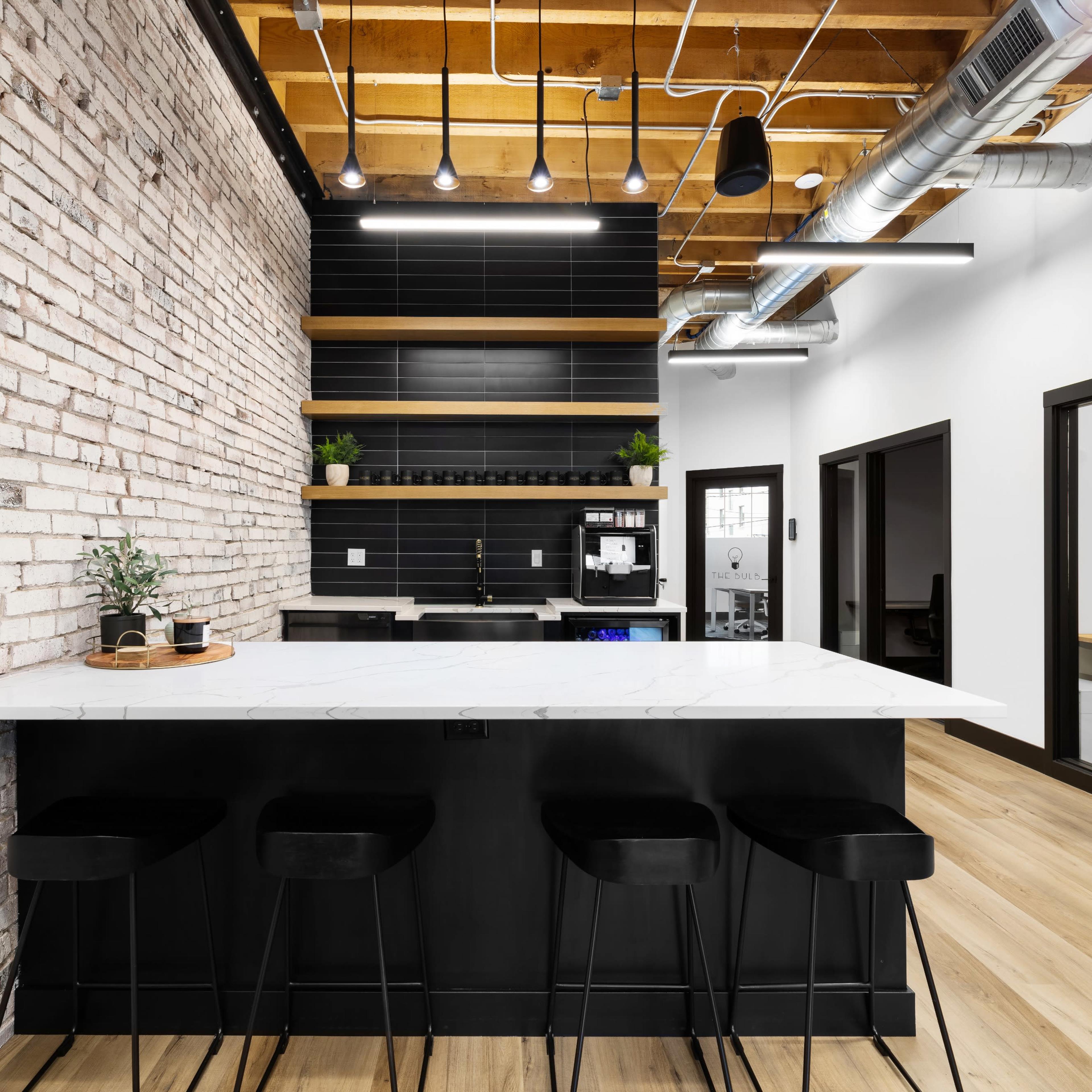 A modern kitchen area features a white marble countertop with black stools, against a backdrop of exposed brick walls and sleek black shelving.