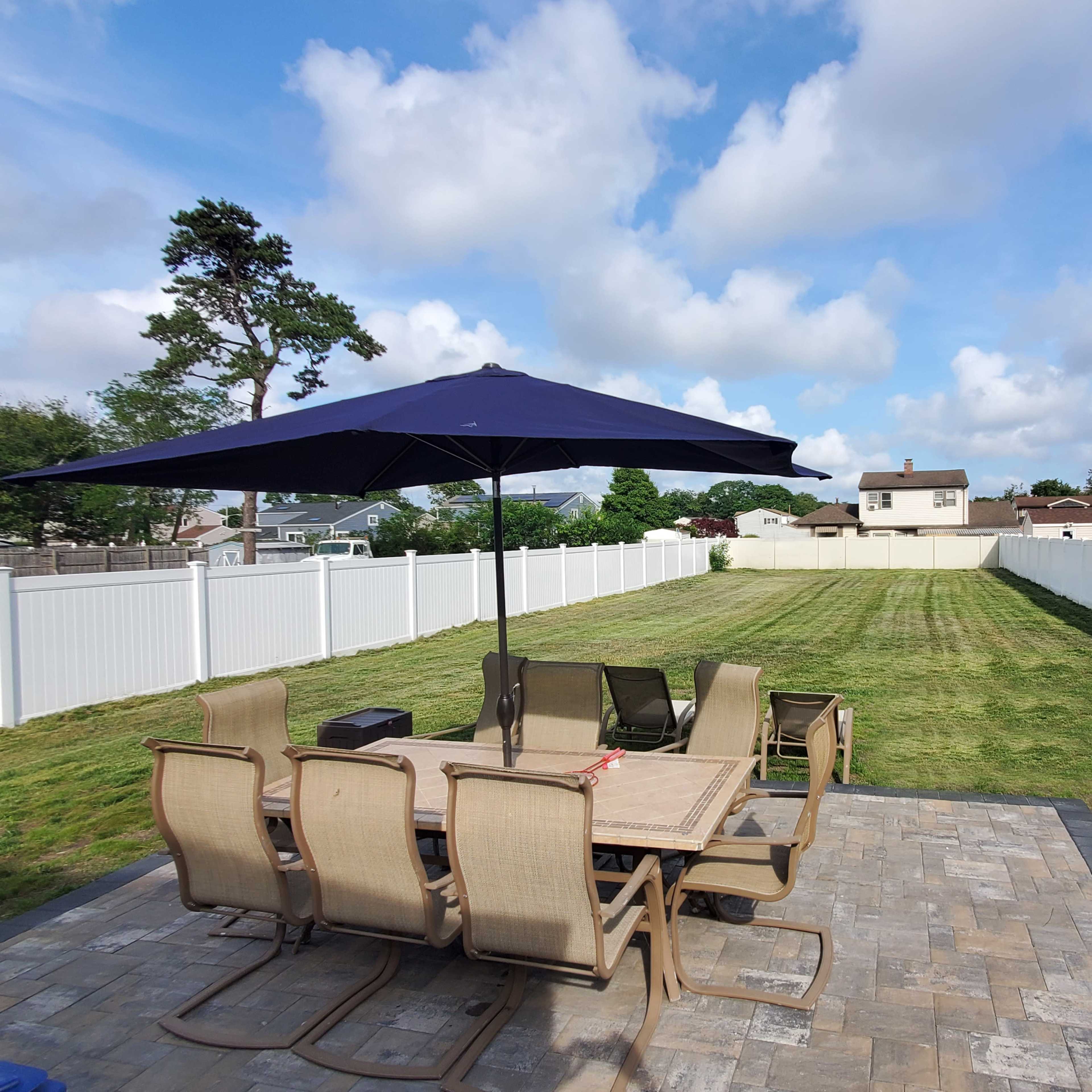 A patio set with a large umbrella overlooks a neatly mowed backyard enclosed by a white picket fence.