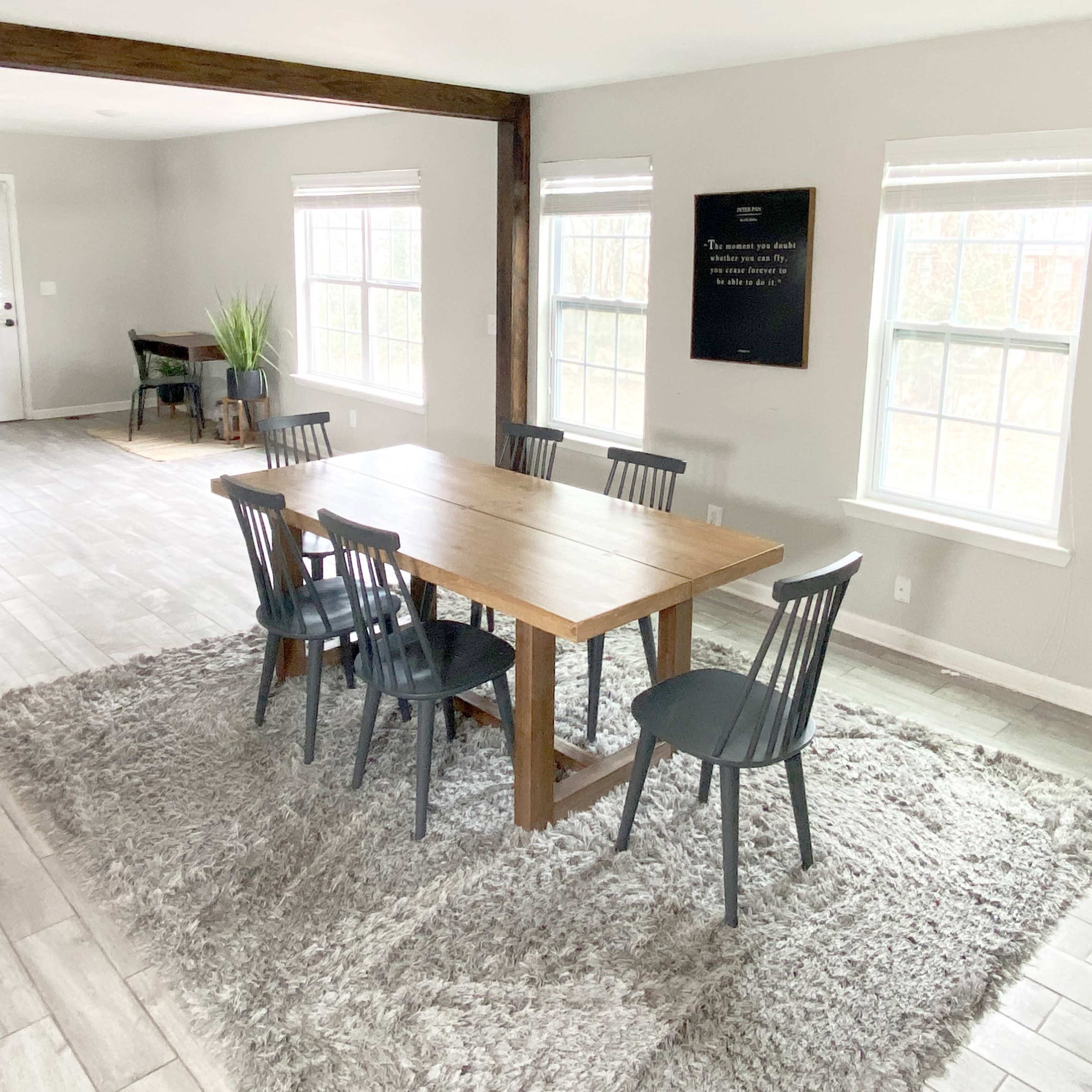 A dining area features a wooden table surrounded by chairs on a plush rug, with large windows letting in natural light.