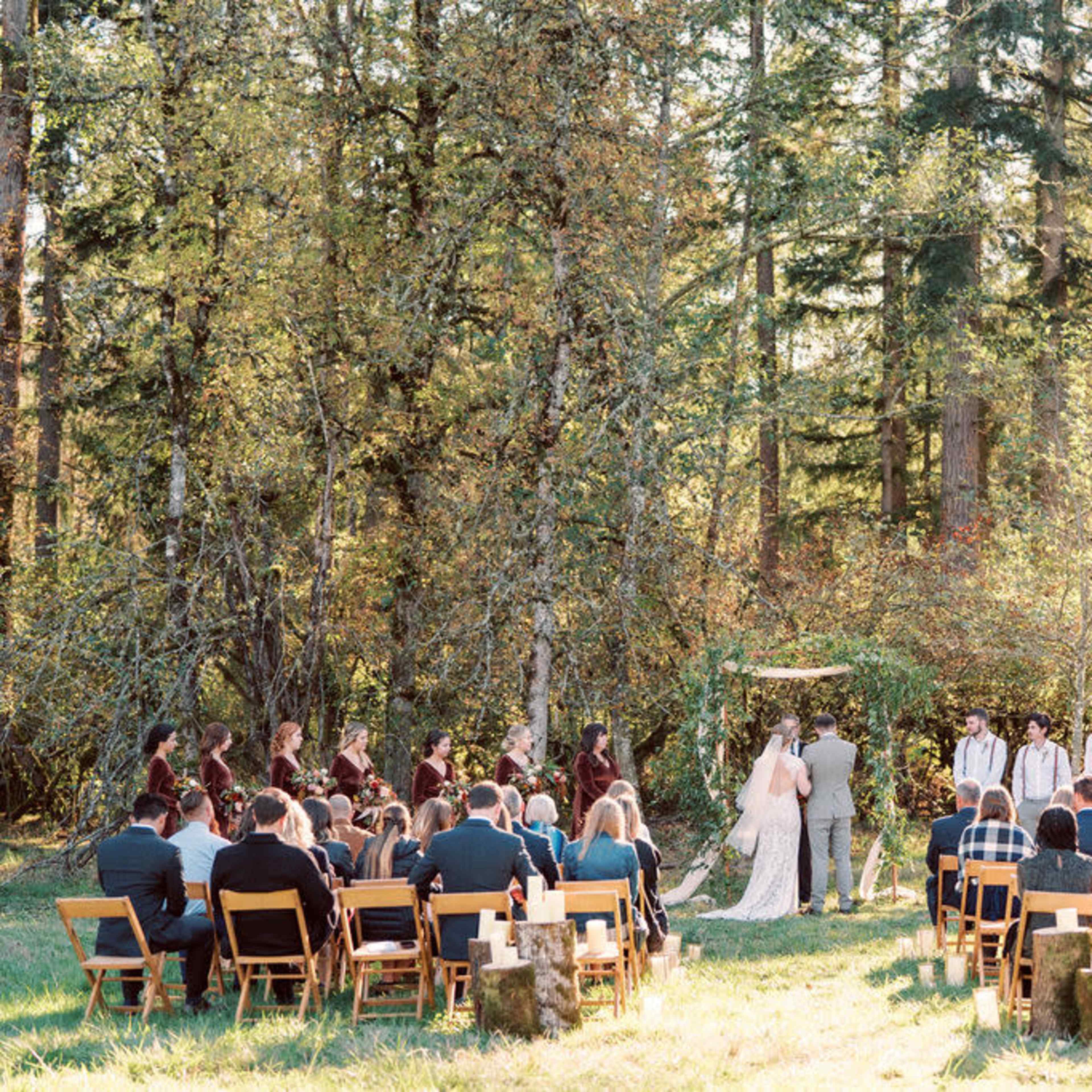 A wedding ceremony takes place outdoors in a forest setting, with a couple in front of an altar and guests seated on chairs arranged in a semicircle.