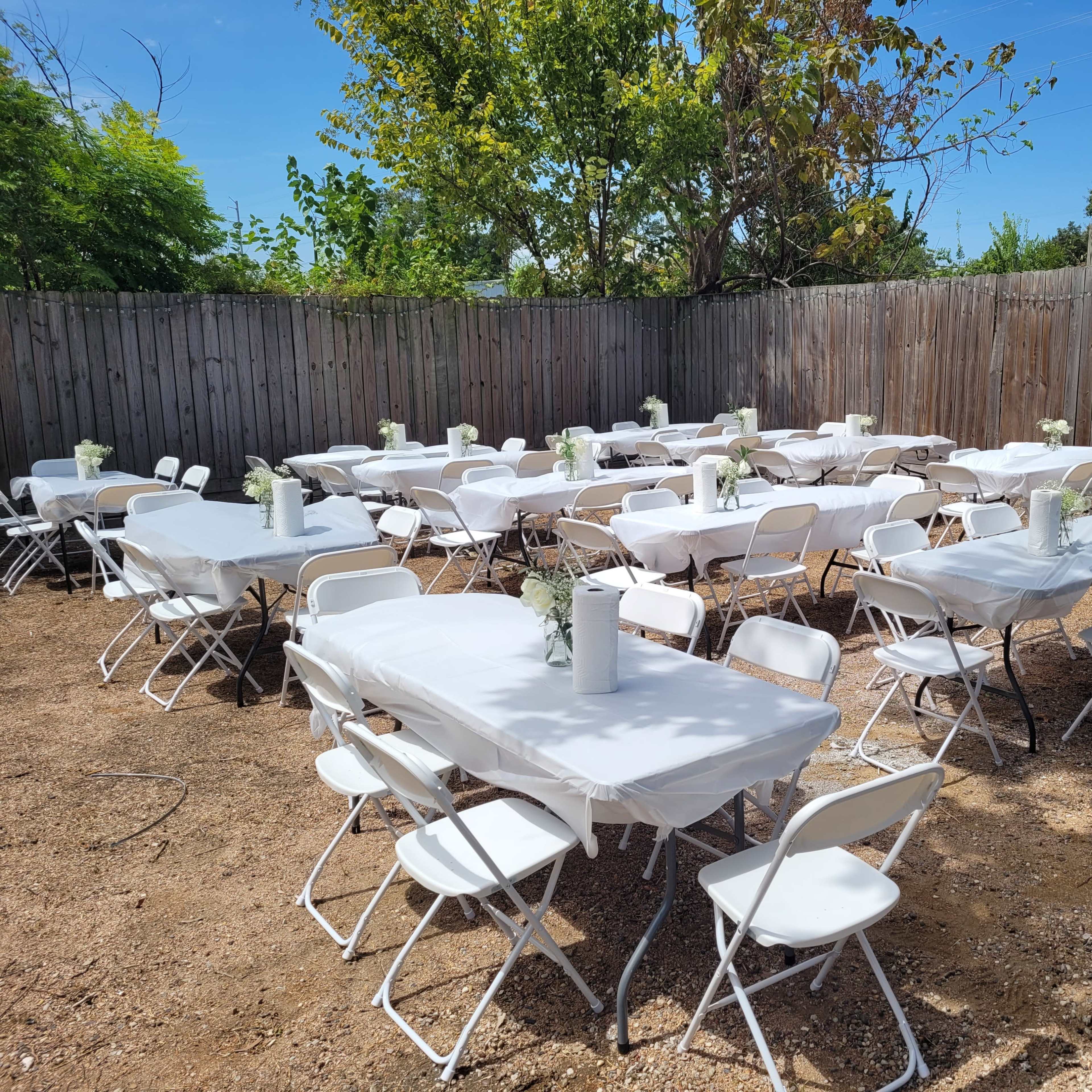 A fenced outdoor space features multiple white tables and chairs arranged for an event, with small centerpieces on each table.