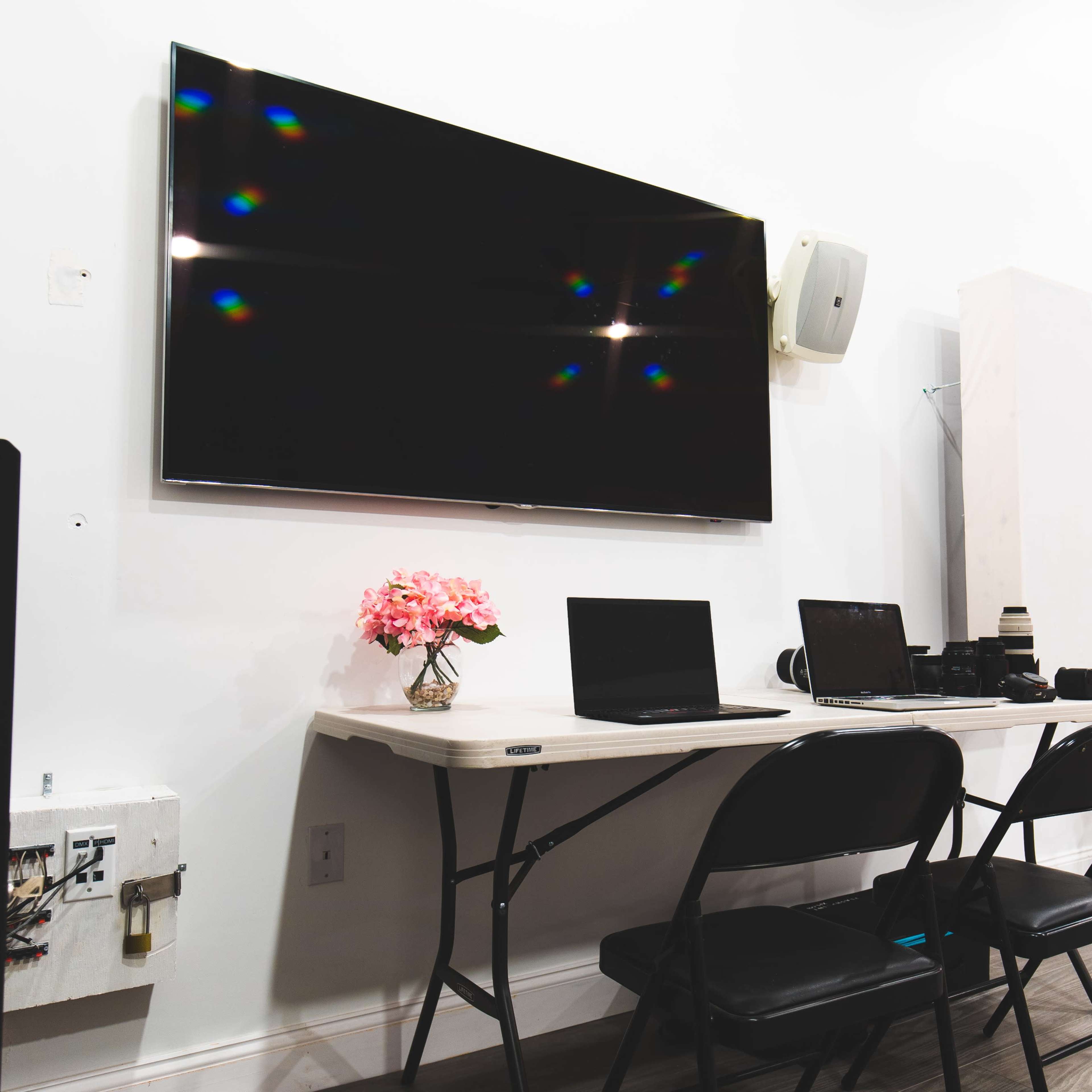 A modern workspace features a table with two laptops, a pink flower arrangement, and a large wall-mounted TV.