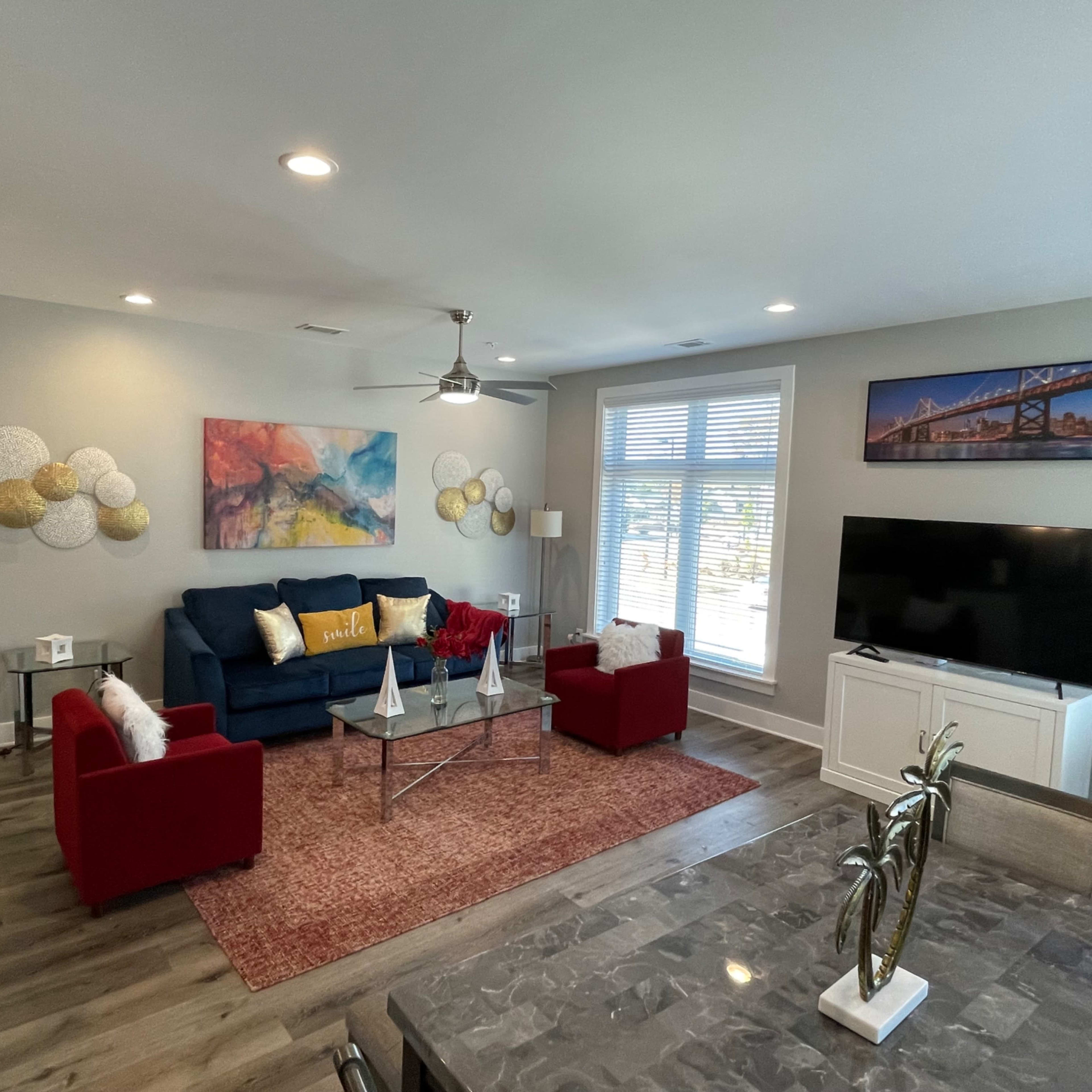 A modern living room with a blue sofa, red chairs, a coffee table, and a large television, all illuminated by natural light from a nearby window.