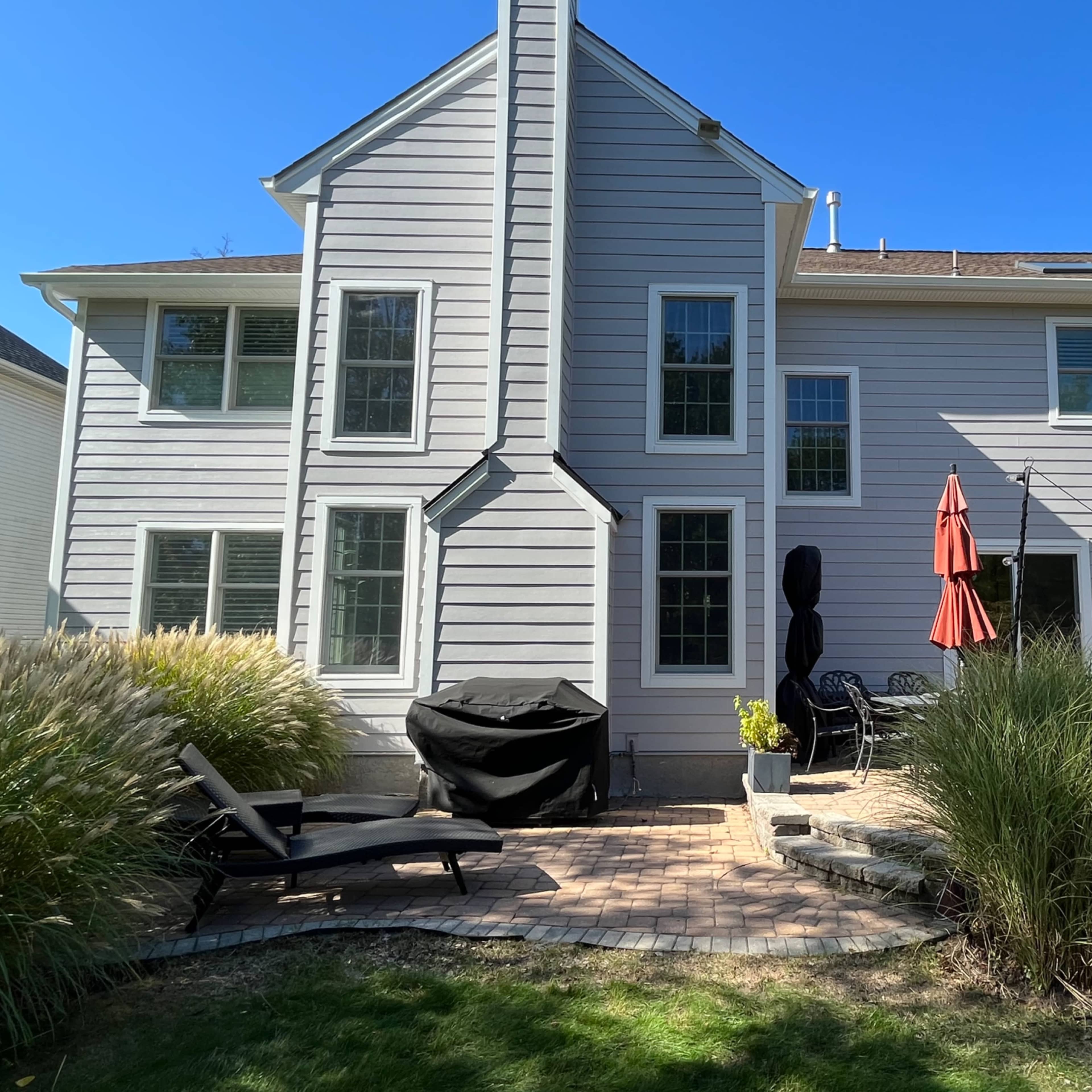 The back of a two-story gray house with several windows, a patio area featuring a grill and seating, and two red umbrellas.
