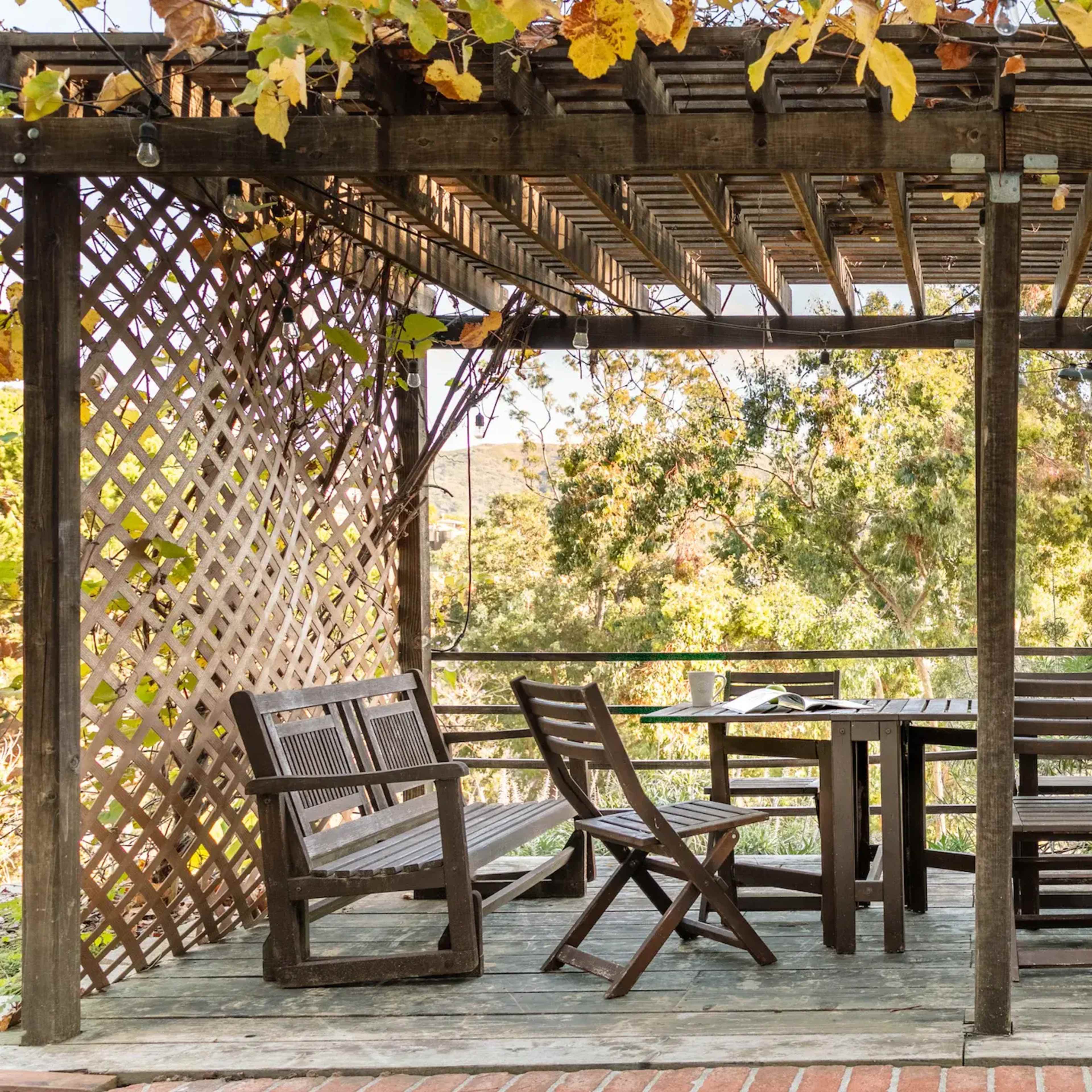 A wooden gazebo with a slatted roof and a table surrounded by chairs overlooks a landscape filled with trees.