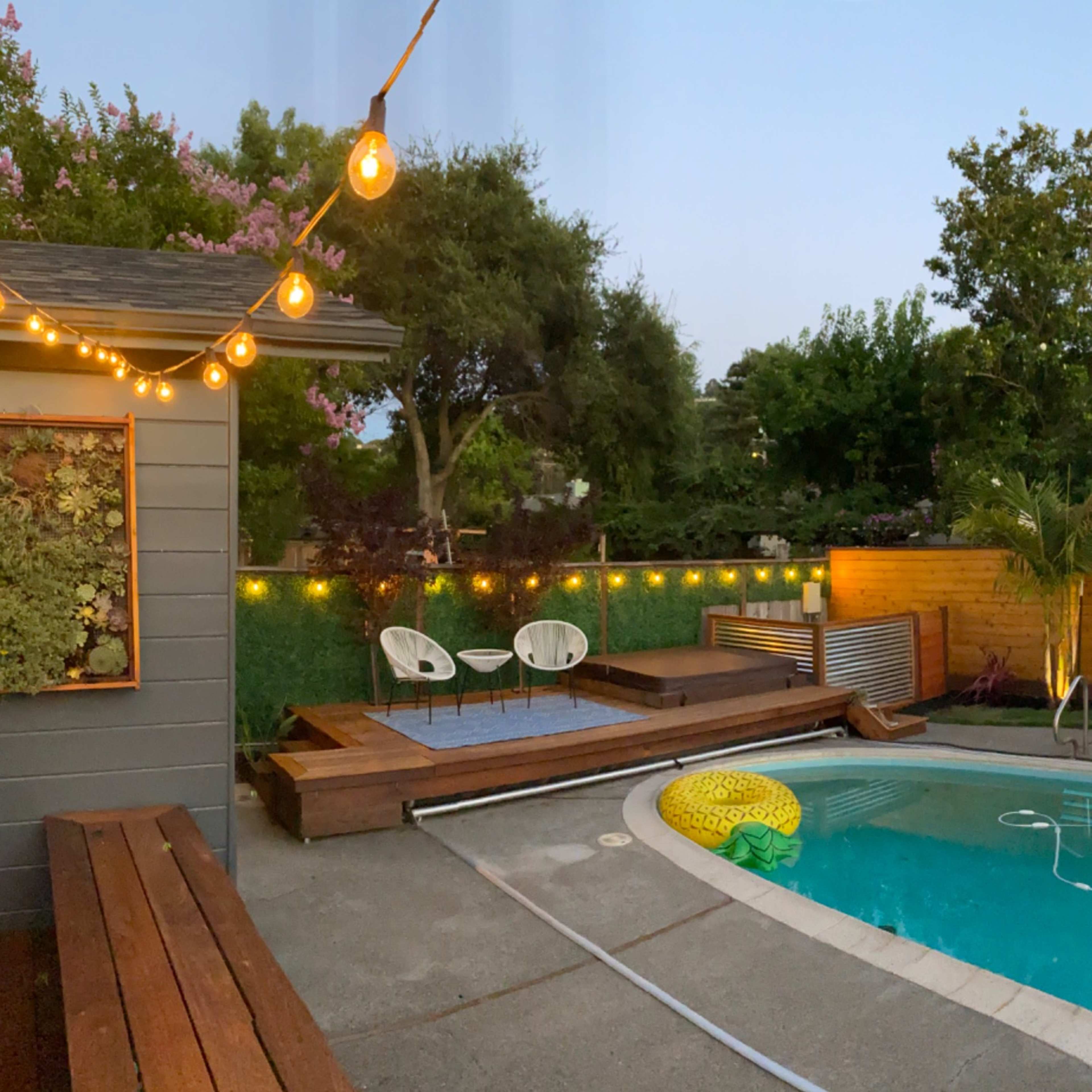 A backyard with a swimming pool, lounge chairs, and string lights illuminating a surrounding landscaped area in the evening.