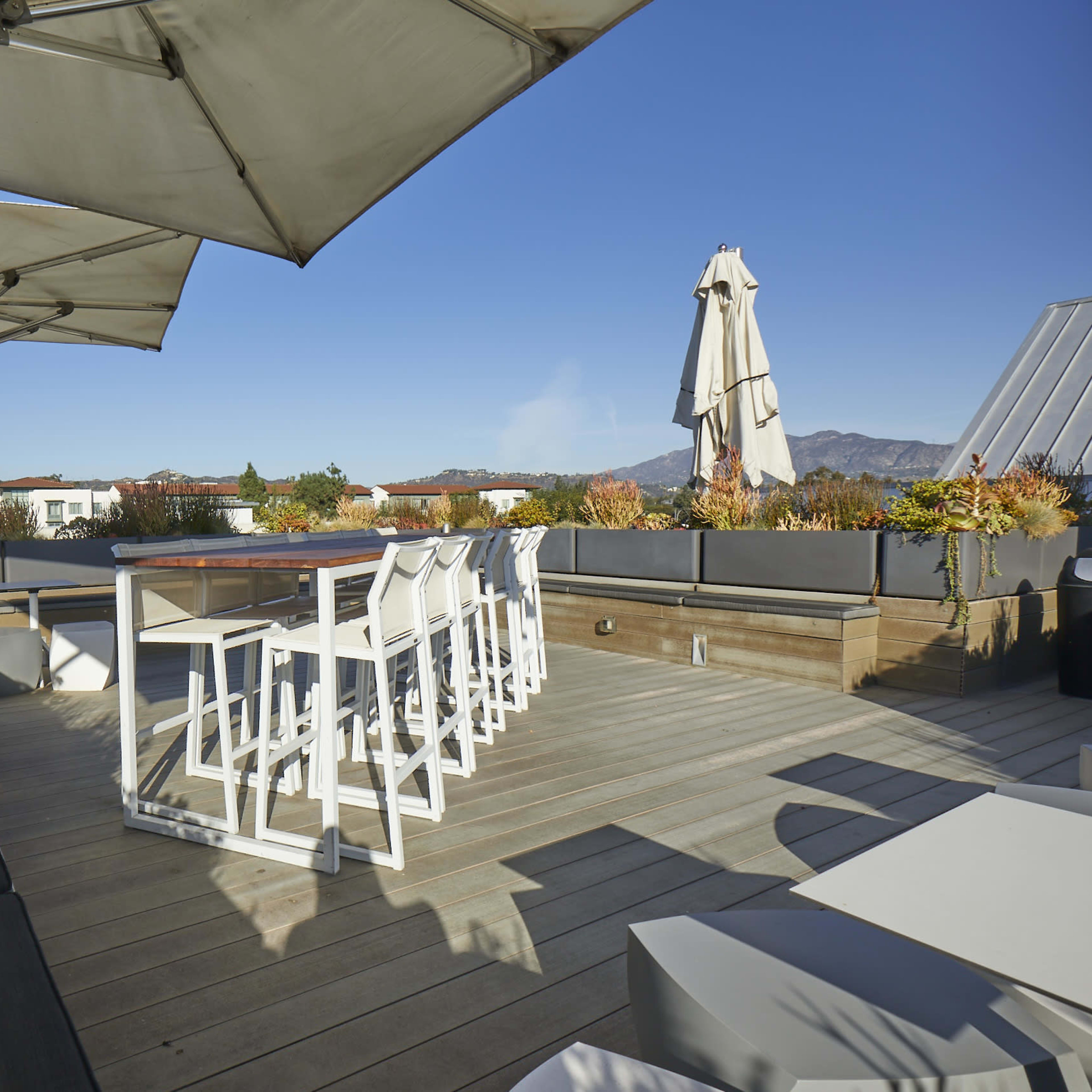 A rooftop patio with seating, large umbrellas, and planters, set against a clear blue sky and distant mountains.