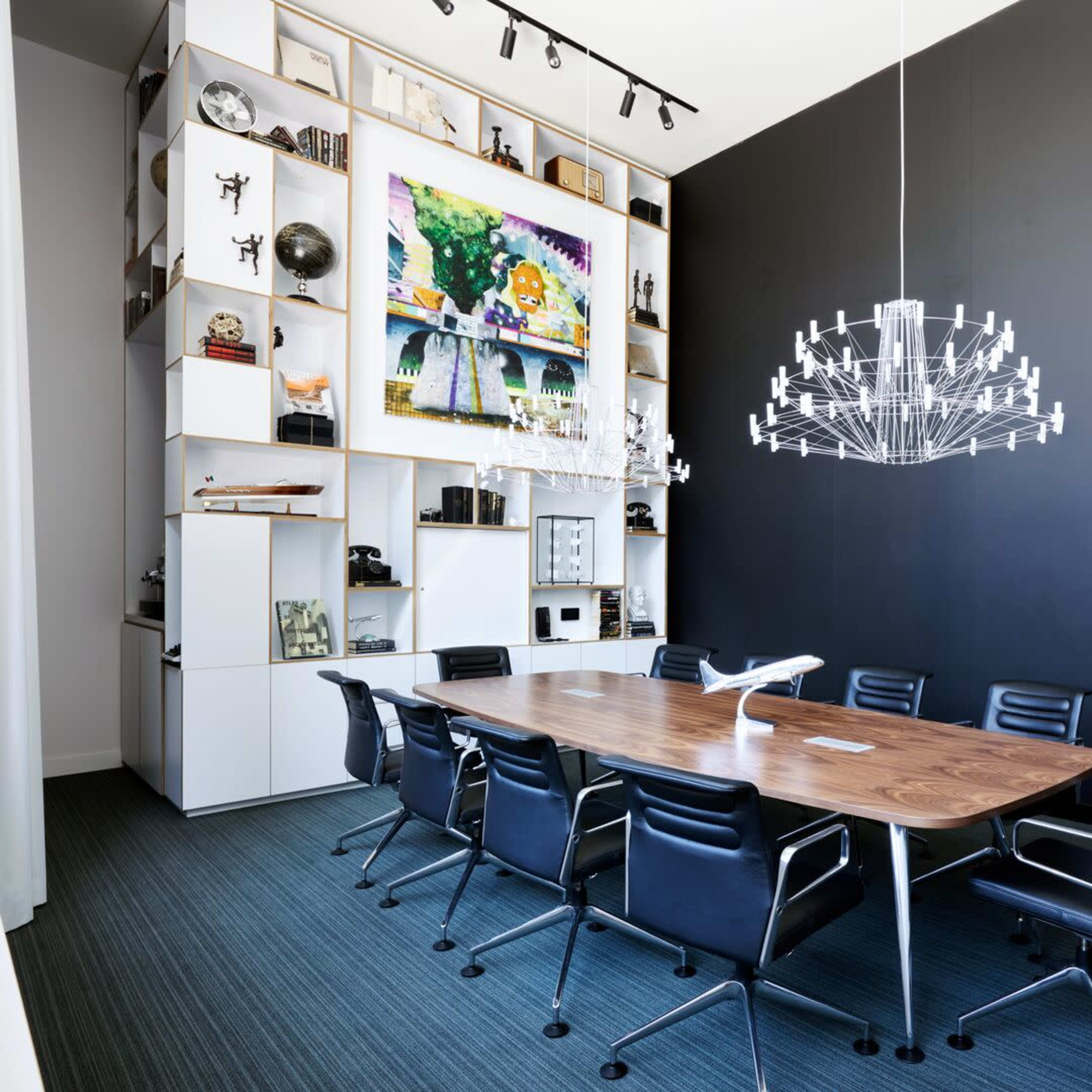 A modern conference room features a large wooden table surrounded by black chairs, with a colorful artwork and shelves displaying various items on the wall.
