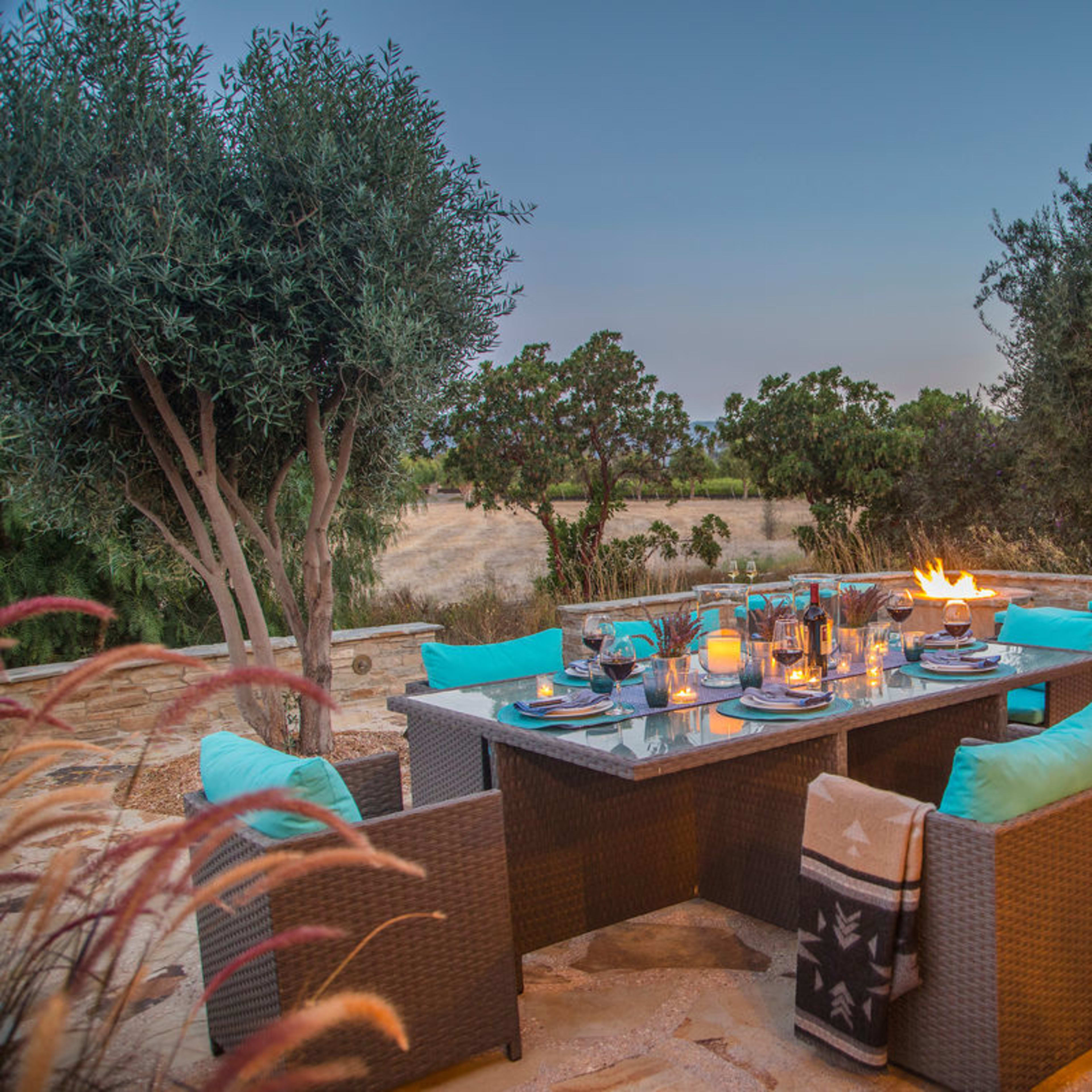 A dining table is set on a stone patio surrounded by trees, with lit candles and blue cushions on the chairs, and a barbecue grill nearby.
