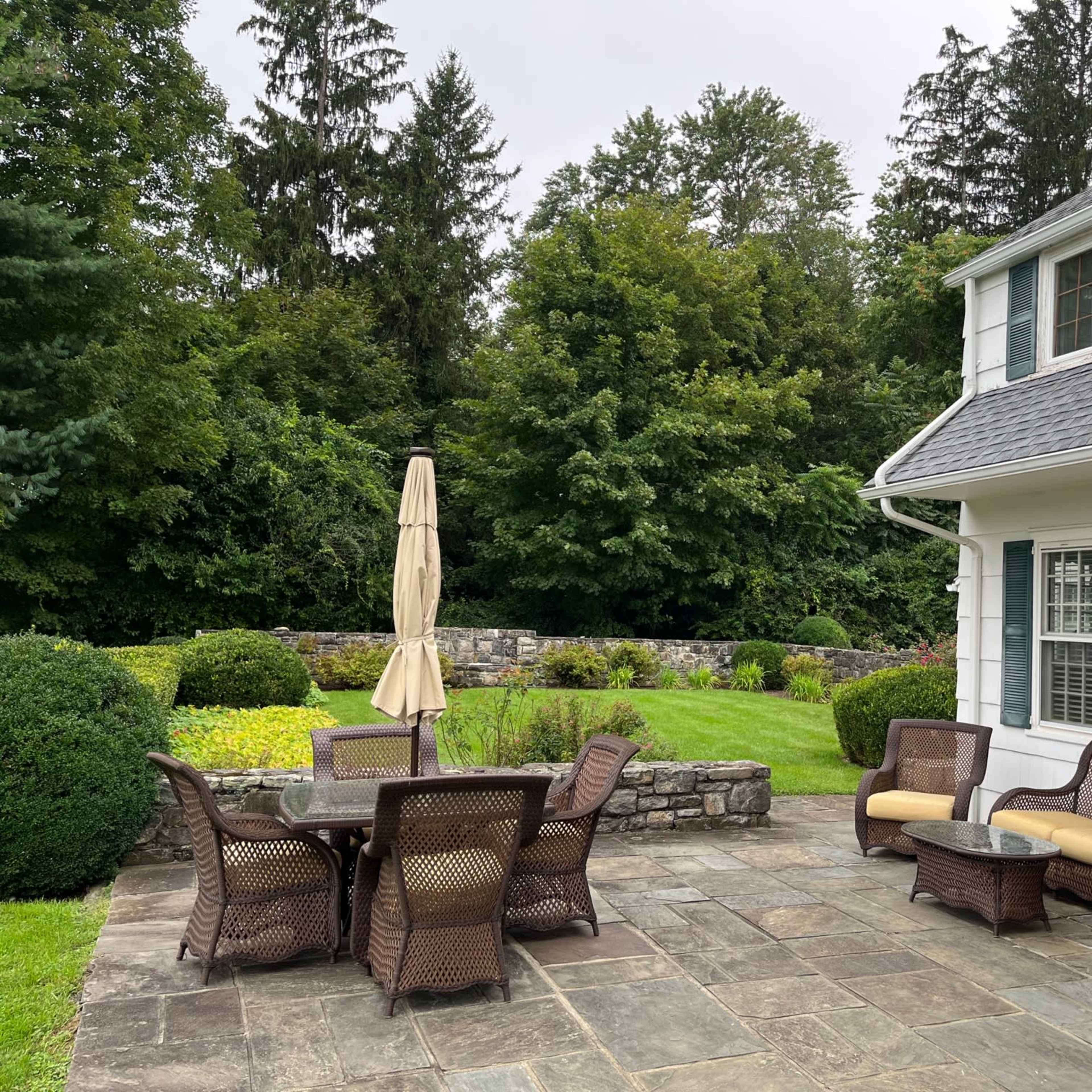 A stone patio with a dining table and chairs, surrounded by a neatly landscaped yard featuring trees and a stone wall.