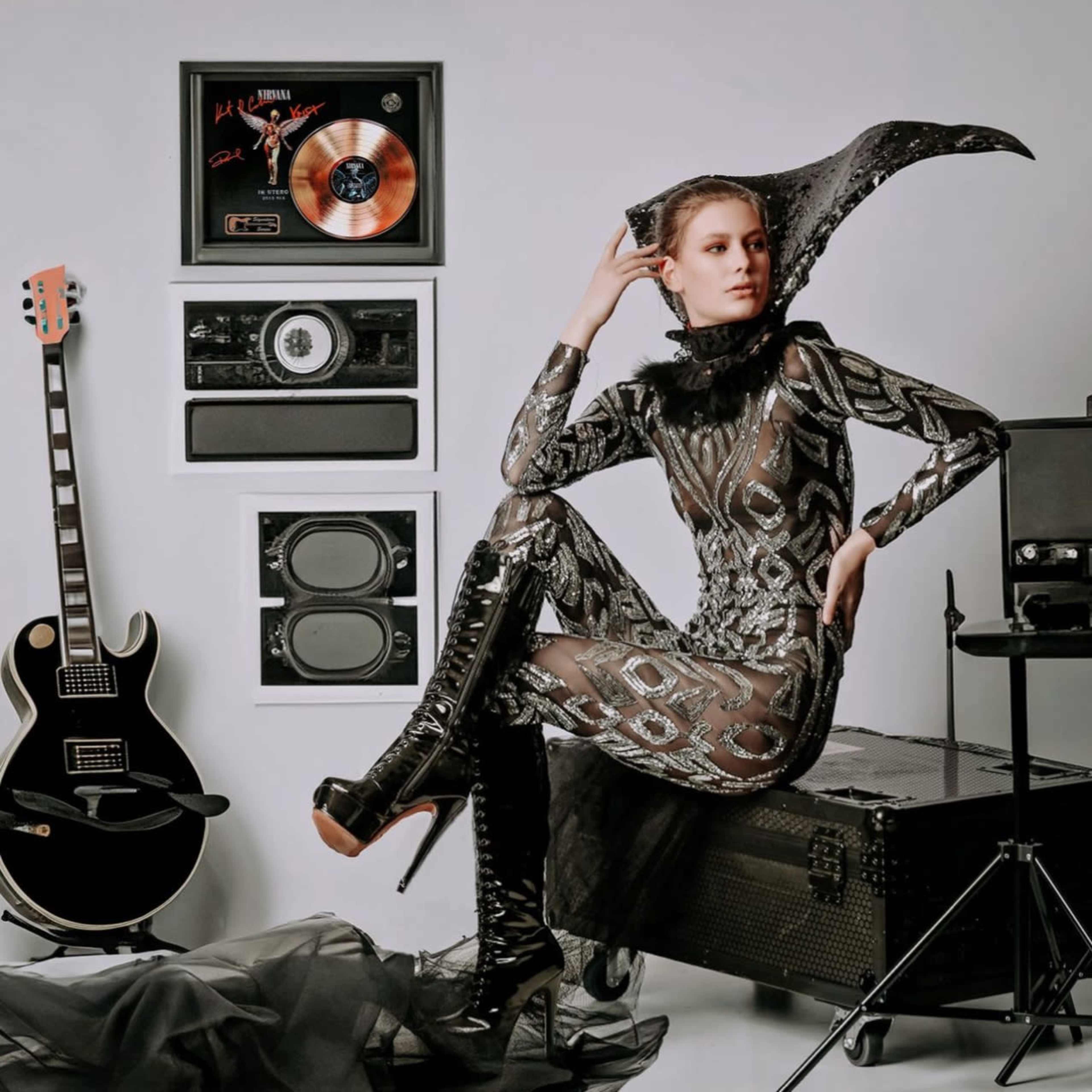 A model in a black, patterned bodysuit and tall heels poses beside musical instruments and framed records against a plain background.