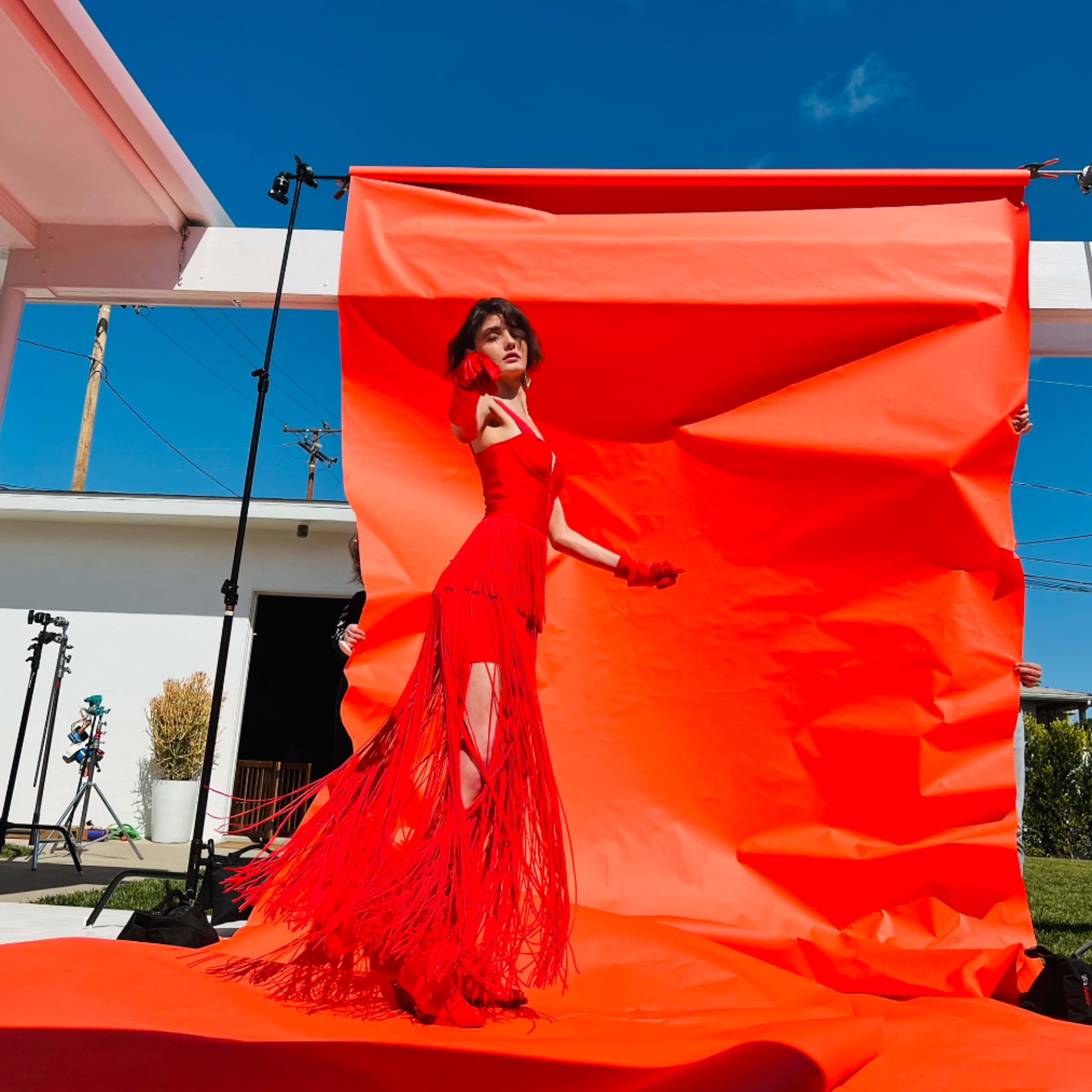 A model poses in a red fringe dress against a bright orange backdrop, with clear blue skies overhead.