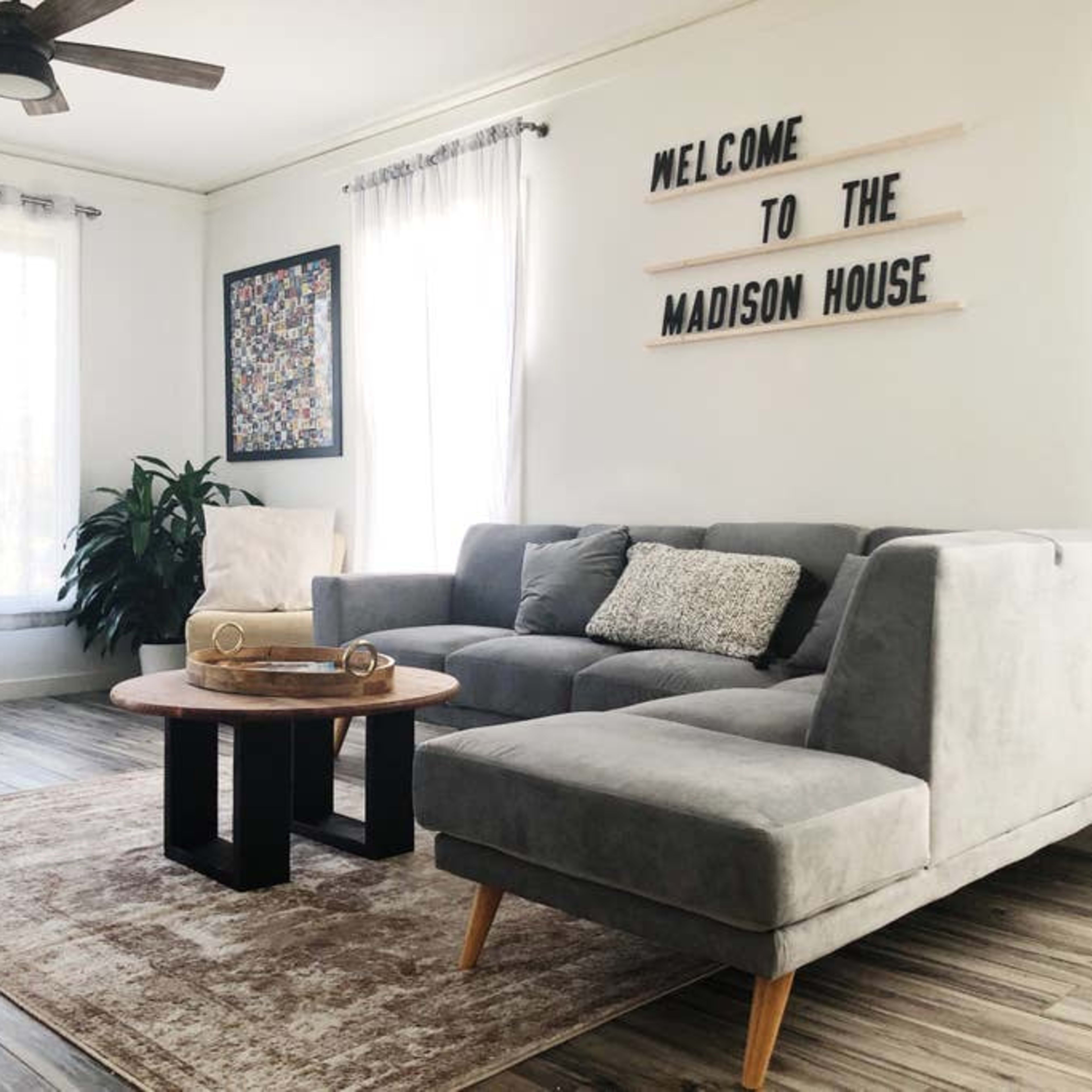 A living room features a gray sectional sofa, a round wooden coffee table, large windows with sheer curtains, and a wall sign that reads "WELCOME TO THE MADISON HOUSE."