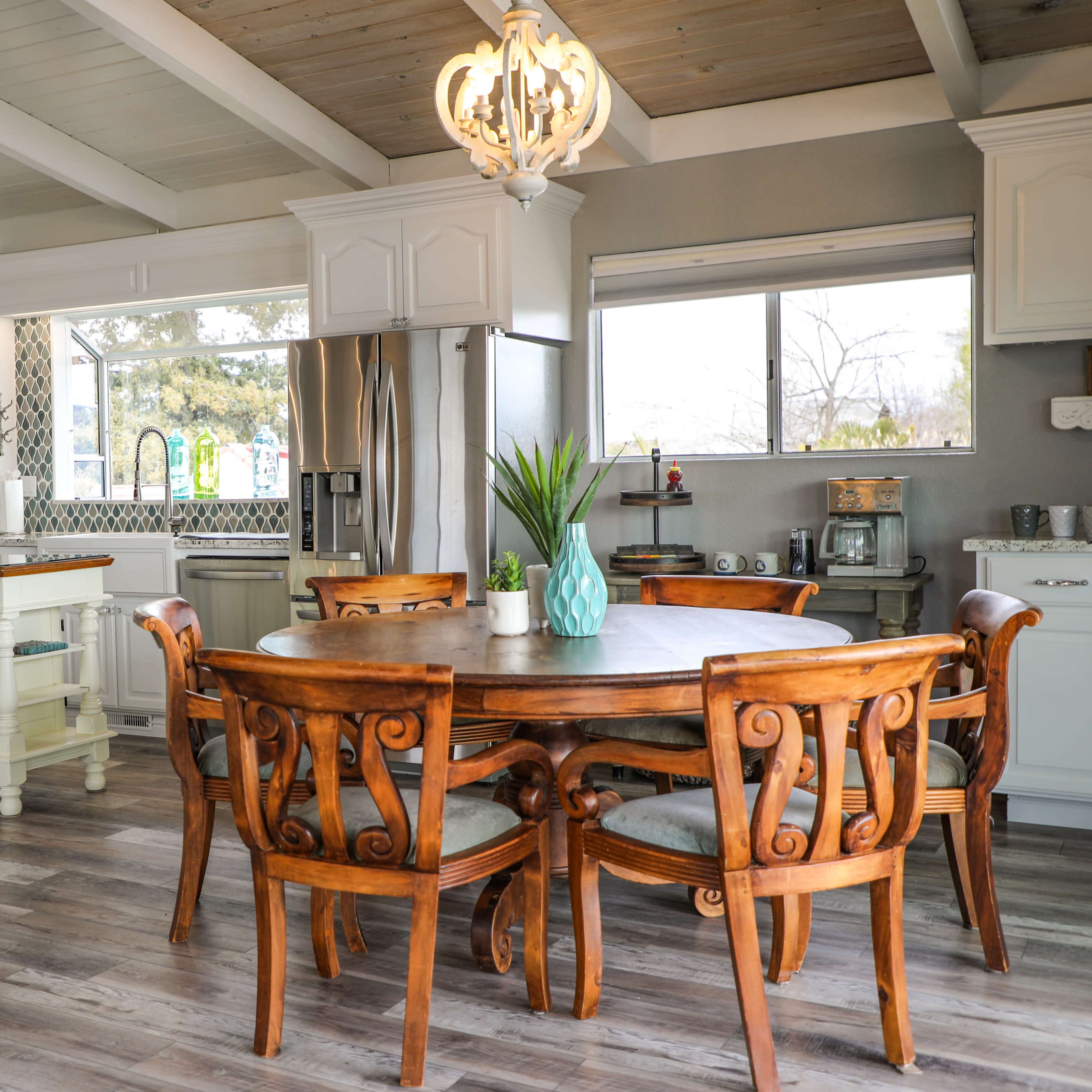 A modern kitchen with a wooden dining table surrounded by ornate chairs, a stainless steel appliance, and a large window letting in natural light.