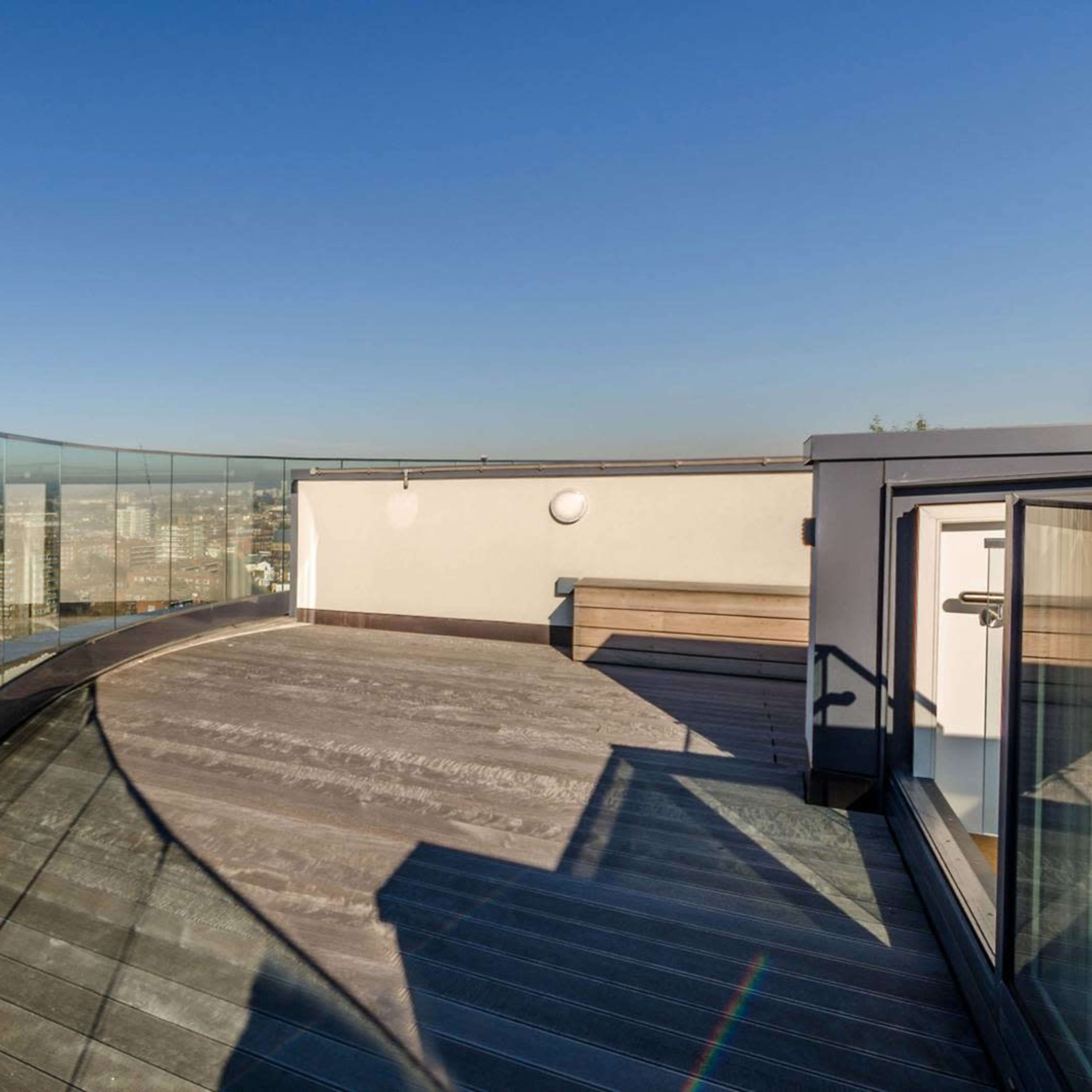 A rooftop terrace with a curved glass railing and wooden flooring, overlooking a cityscape under a clear blue sky.