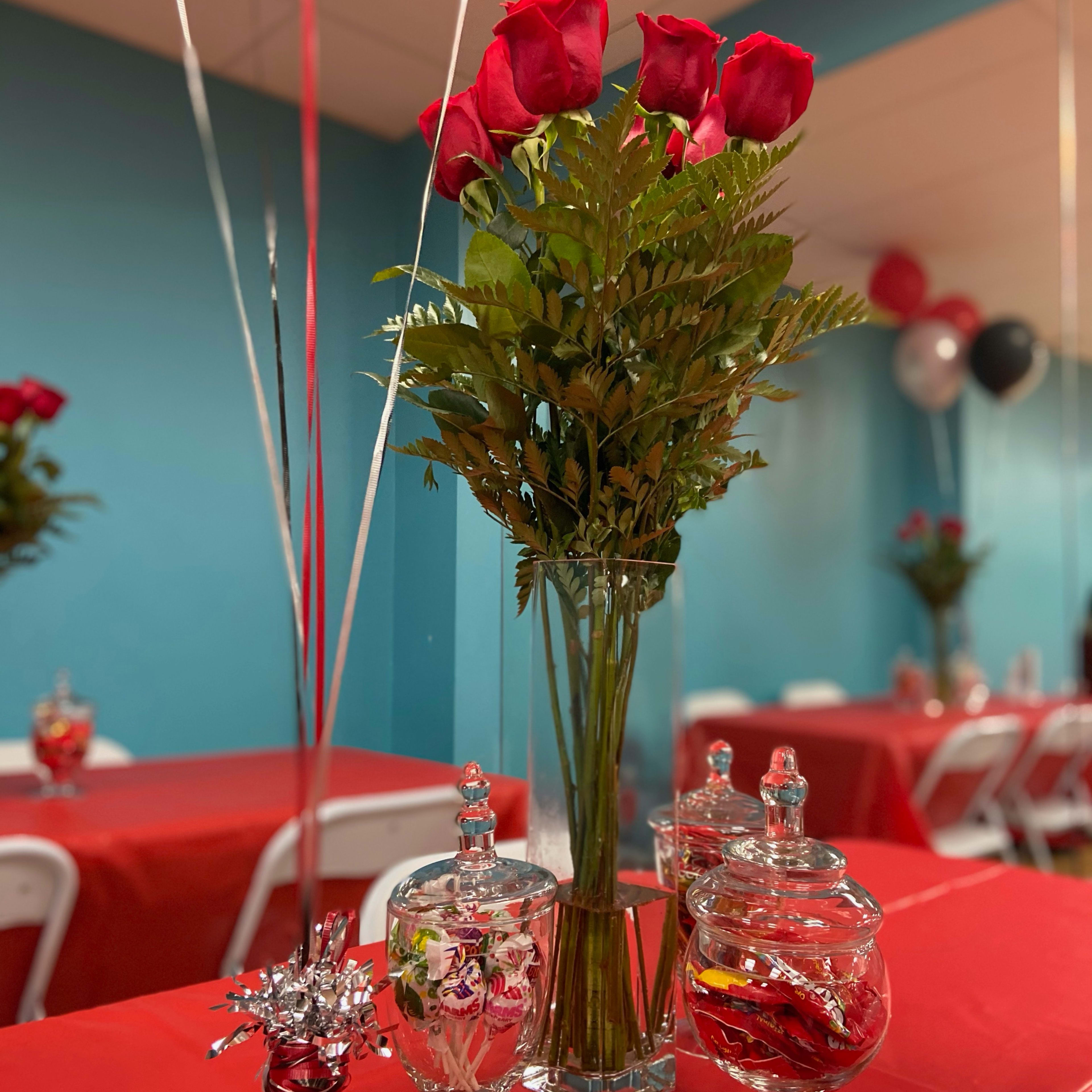 A bouquet of red roses and greenery sits in a clear vase on a table with a red tablecloth, accompanied by decorative glass jars and balloons in a festive setting.