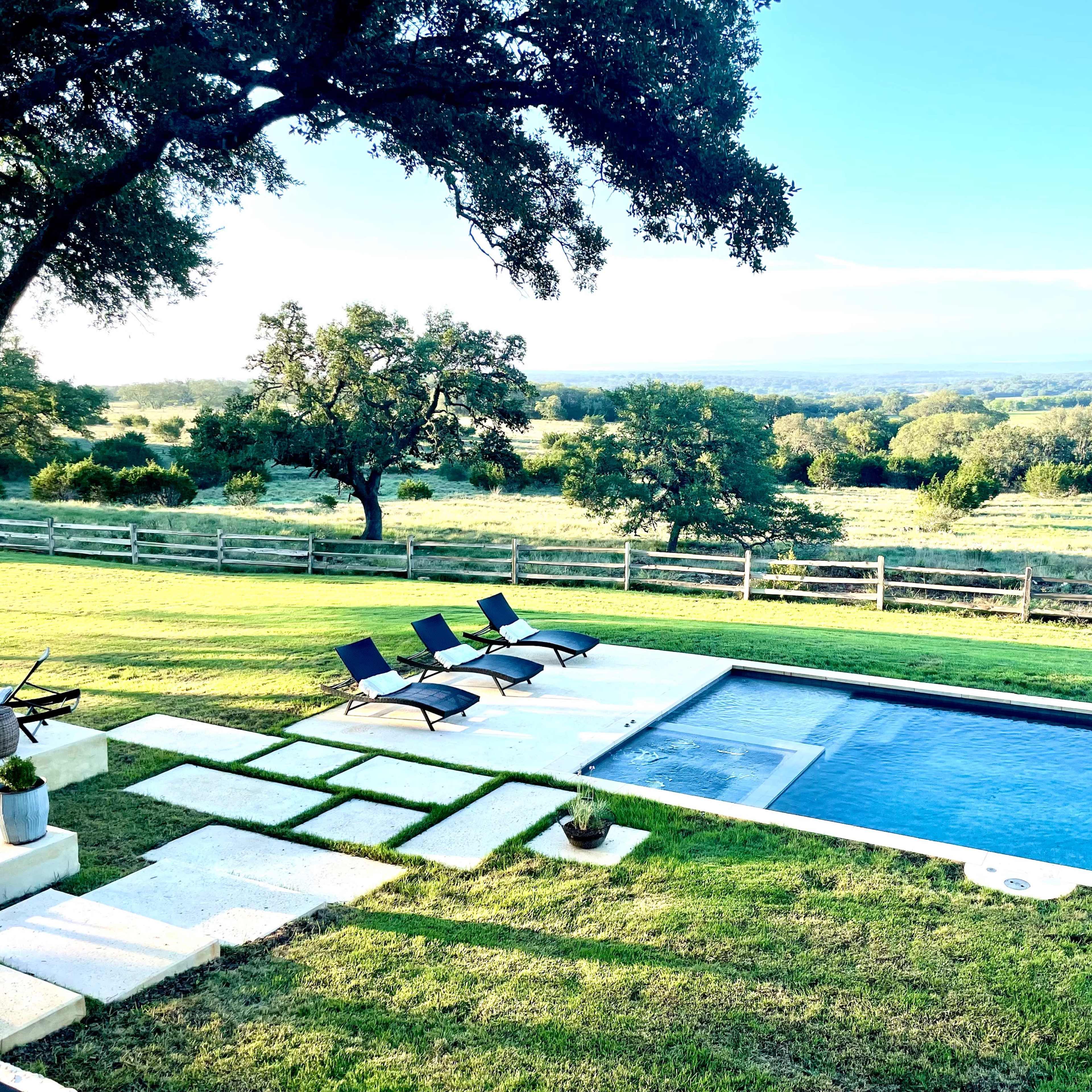 A serene outdoor scene featuring a swimming pool with lounge chairs, surrounded by a landscaped lawn and a scenic view of rolling hills.