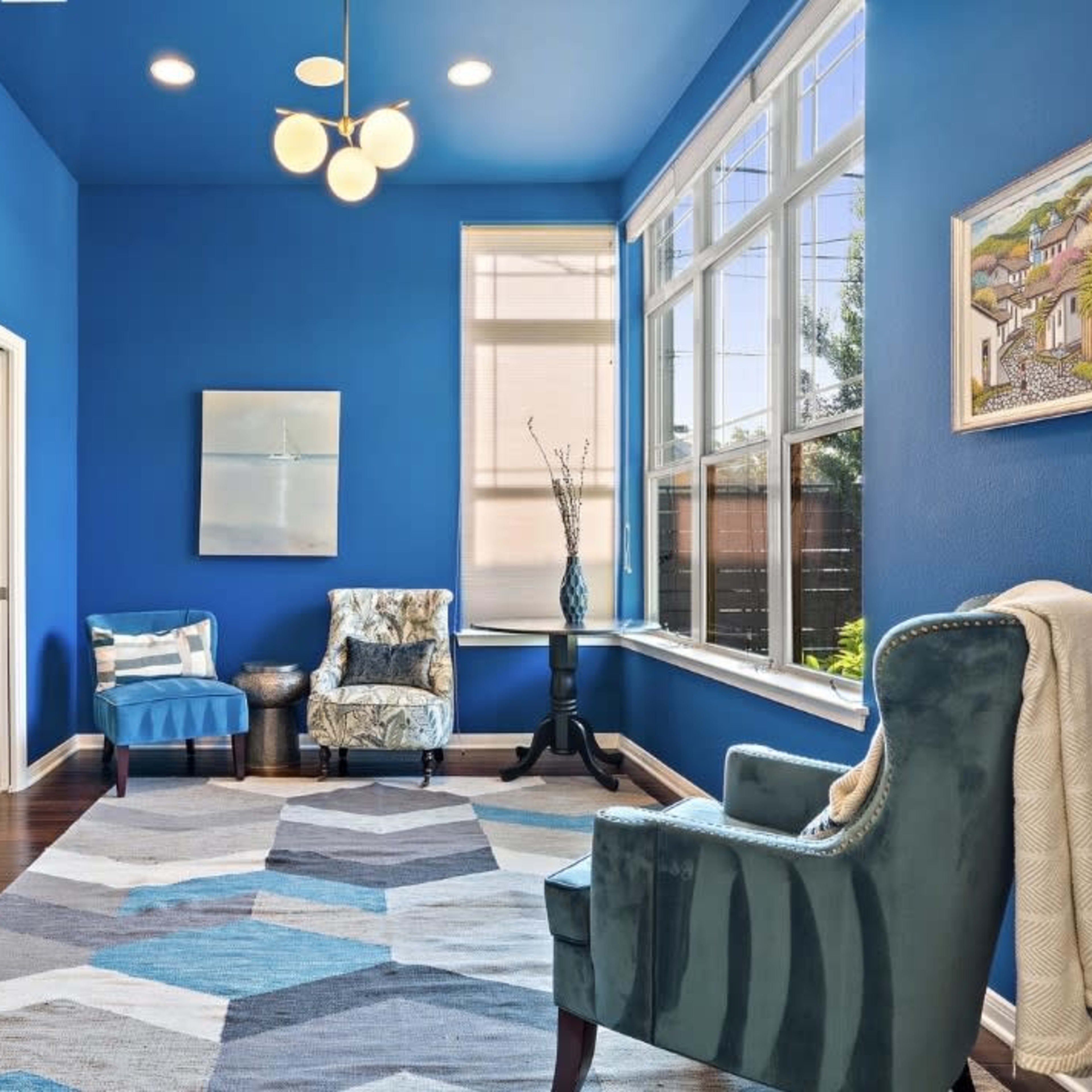 The room features blue walls, a large window, two upholstered chairs, and a geometric patterned rug on a hardwood floor.