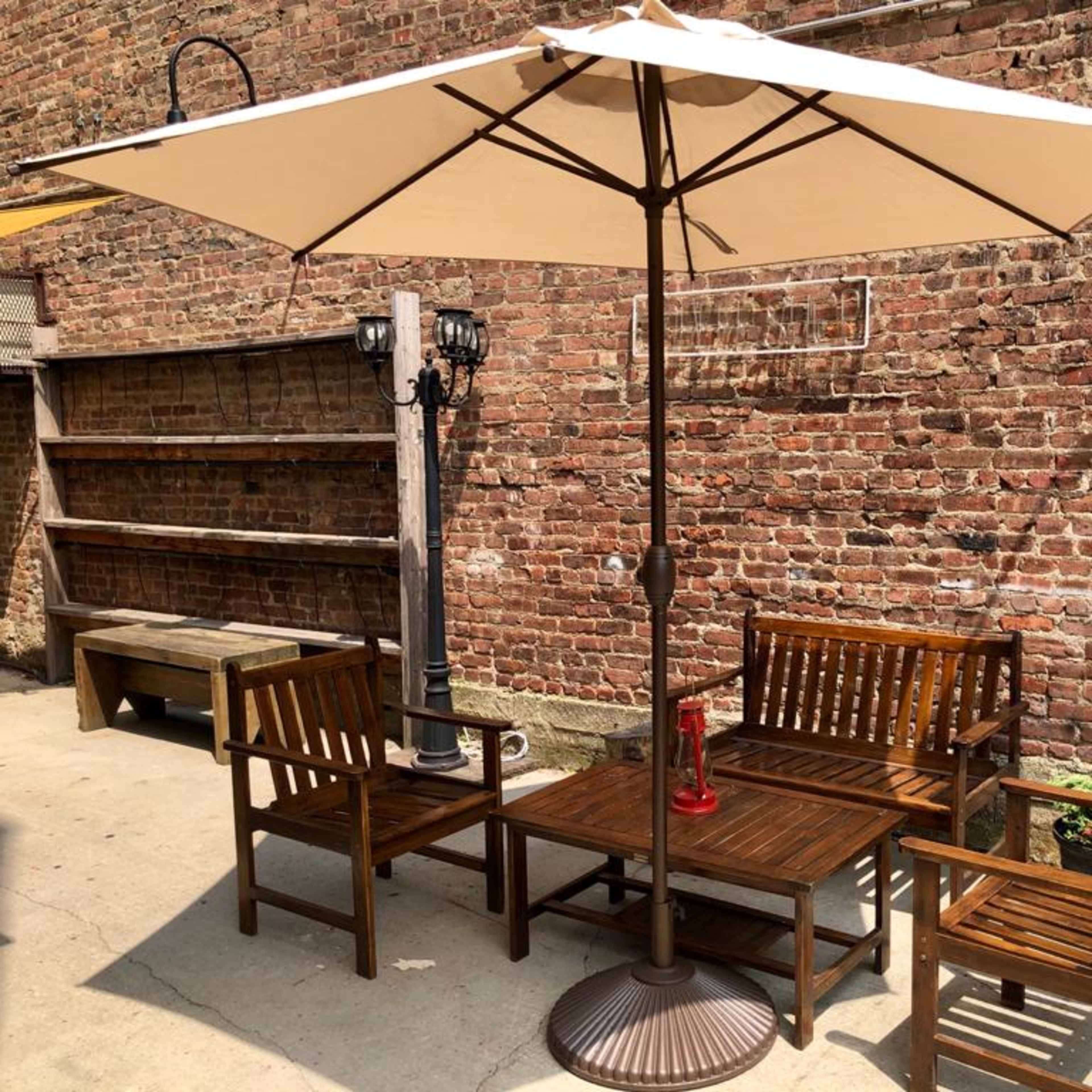 A wooden patio set with an umbrella is set against a brick wall, alongside a small lamp post and a shelf.