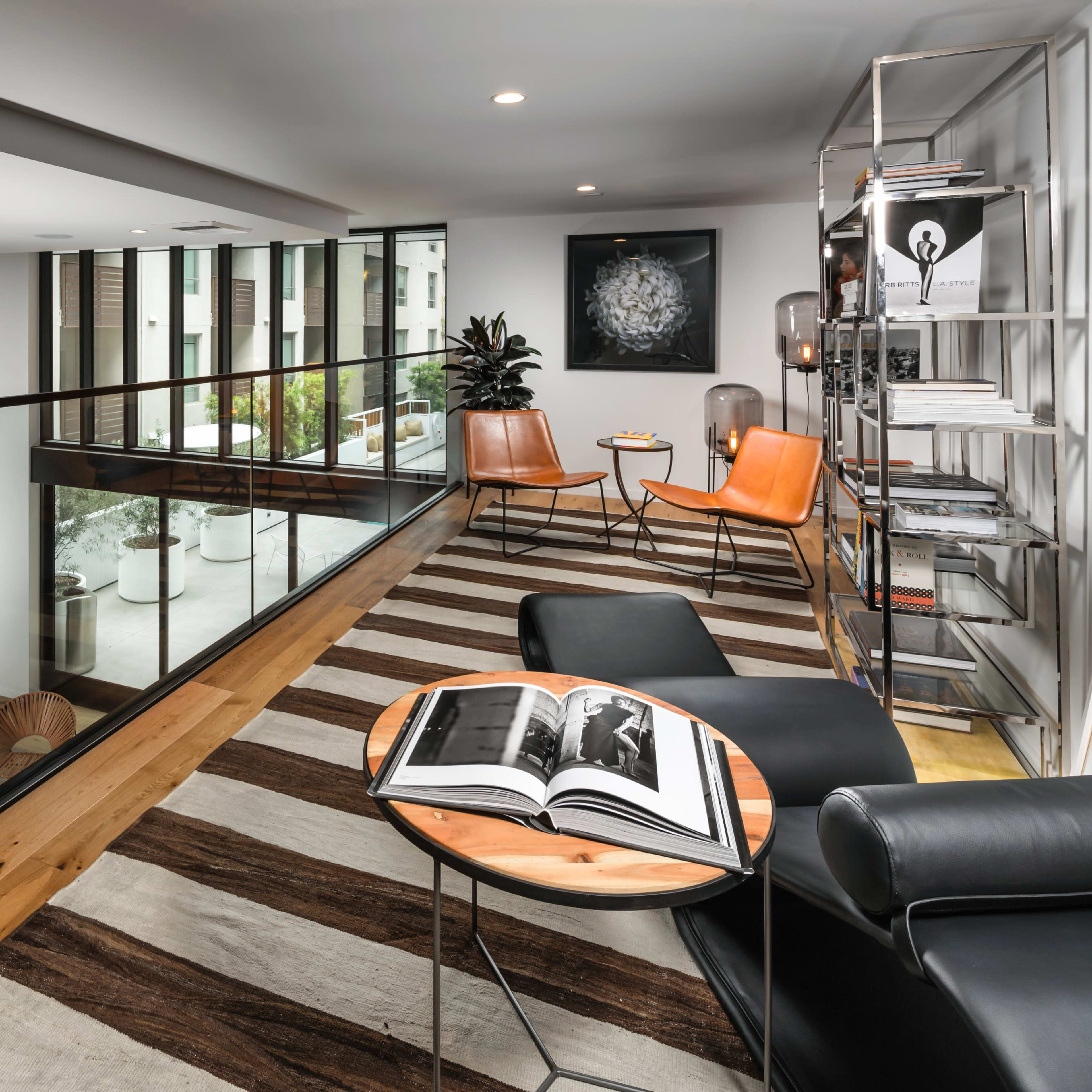 A modern living space with a glass railing balcony overlooking a lower seating area, featuring contemporary furnishings and a striped area rug.