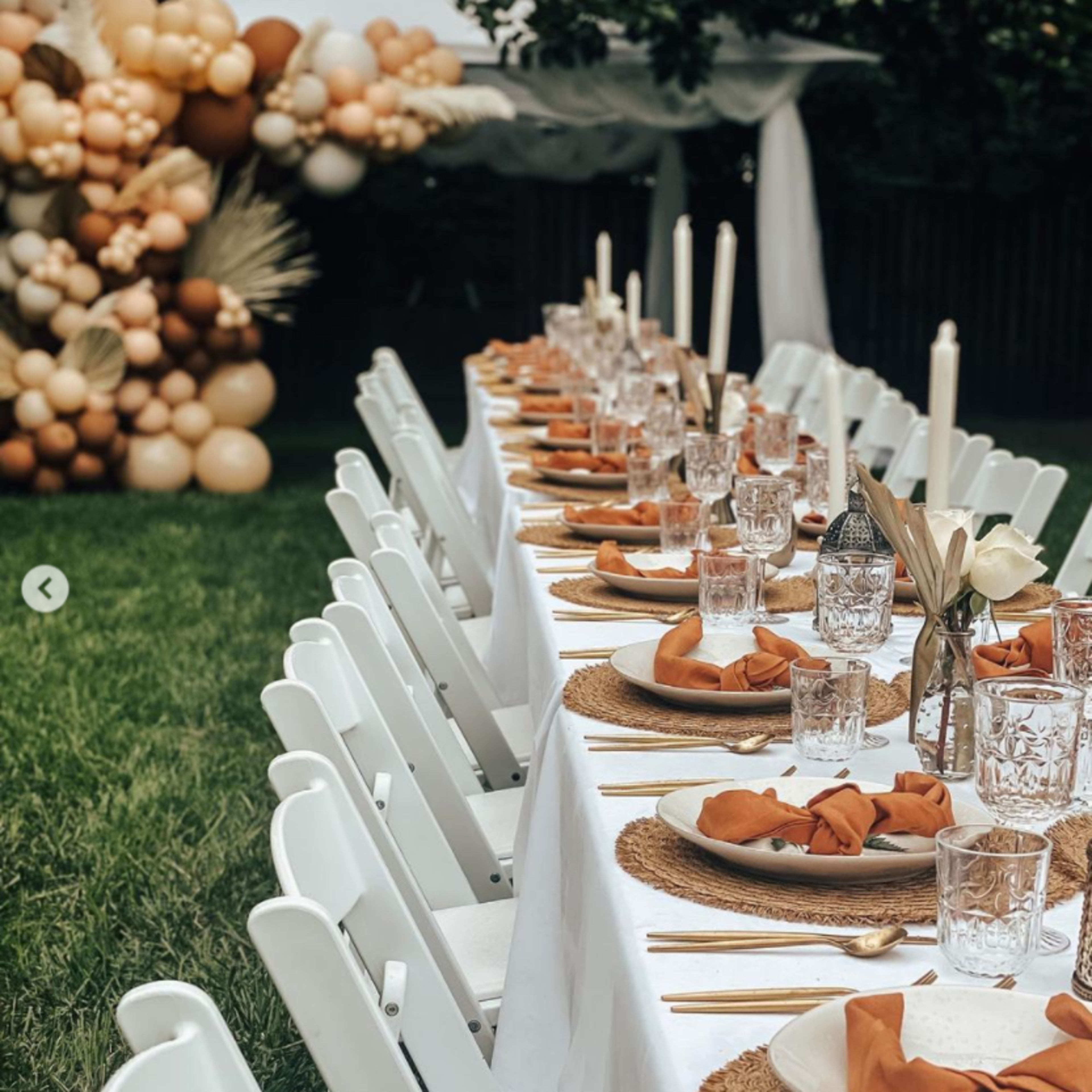 A long dining table is set outdoors with white chairs, decorated with plates, glasses, and orange napkins, in front of a balloon installation.