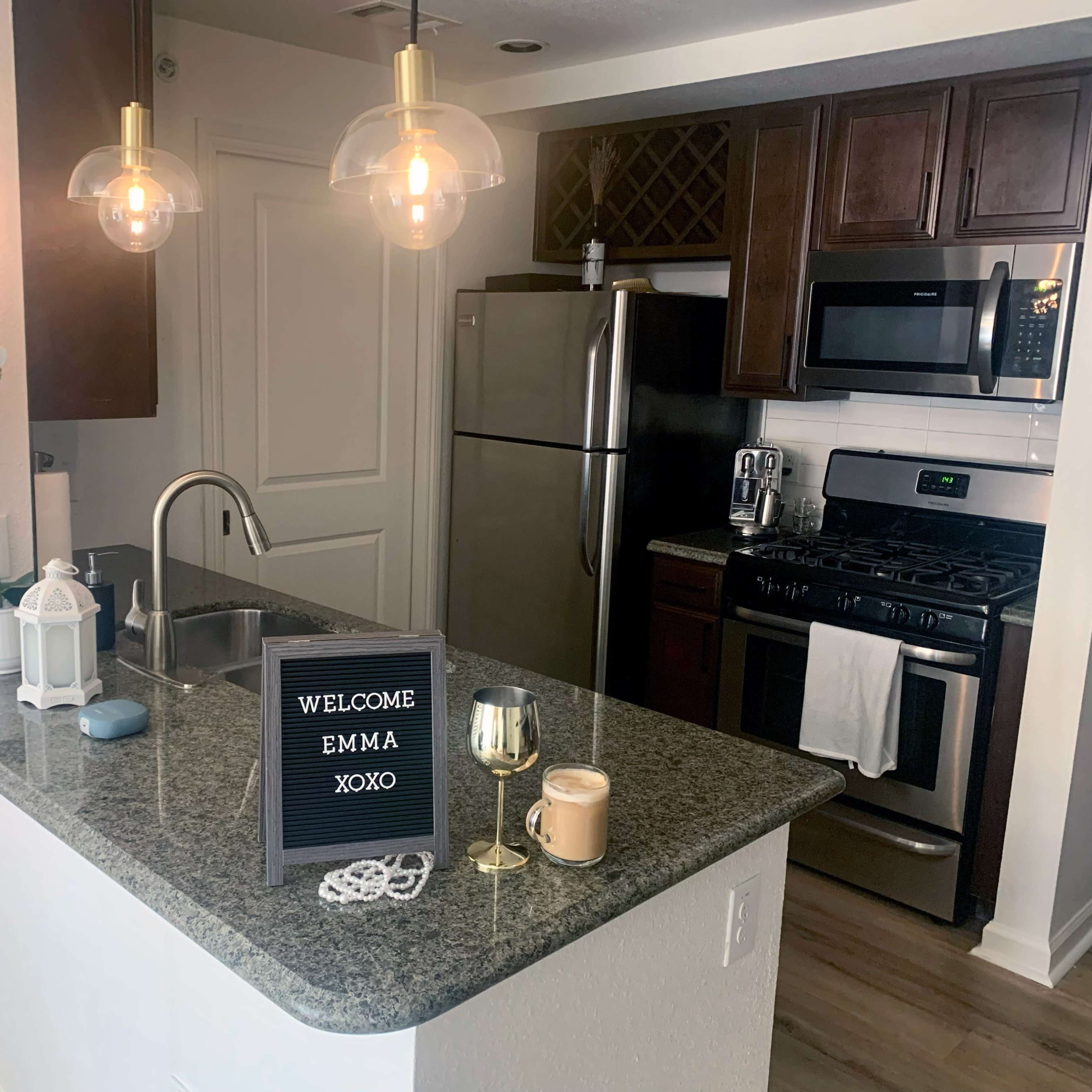 A modern kitchen features a granite countertop, stainless steel appliances, pendant lights, and a welcome sign.