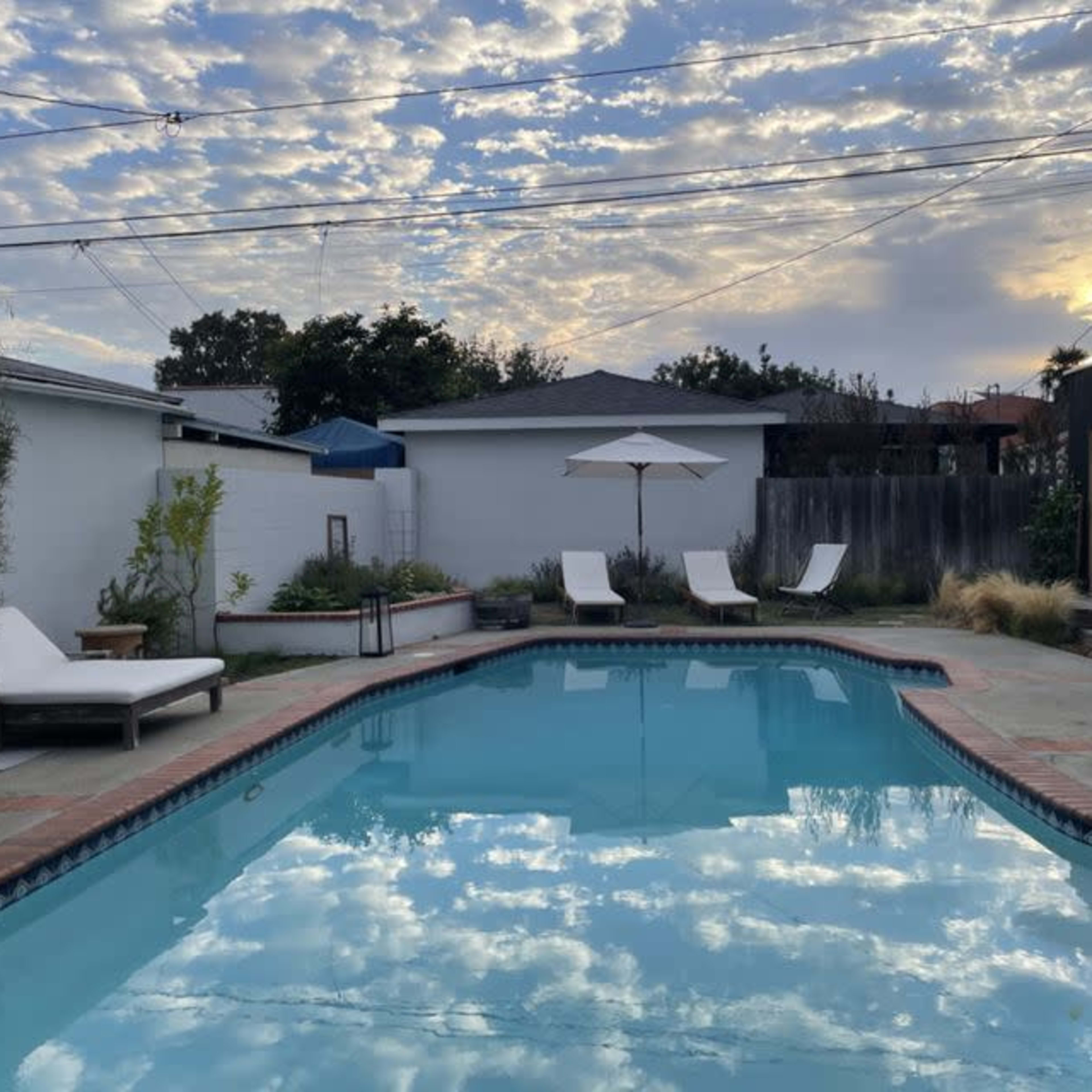 A swimming pool surrounded by lounge chairs and an umbrella, with a cloudy sky reflected in the water.