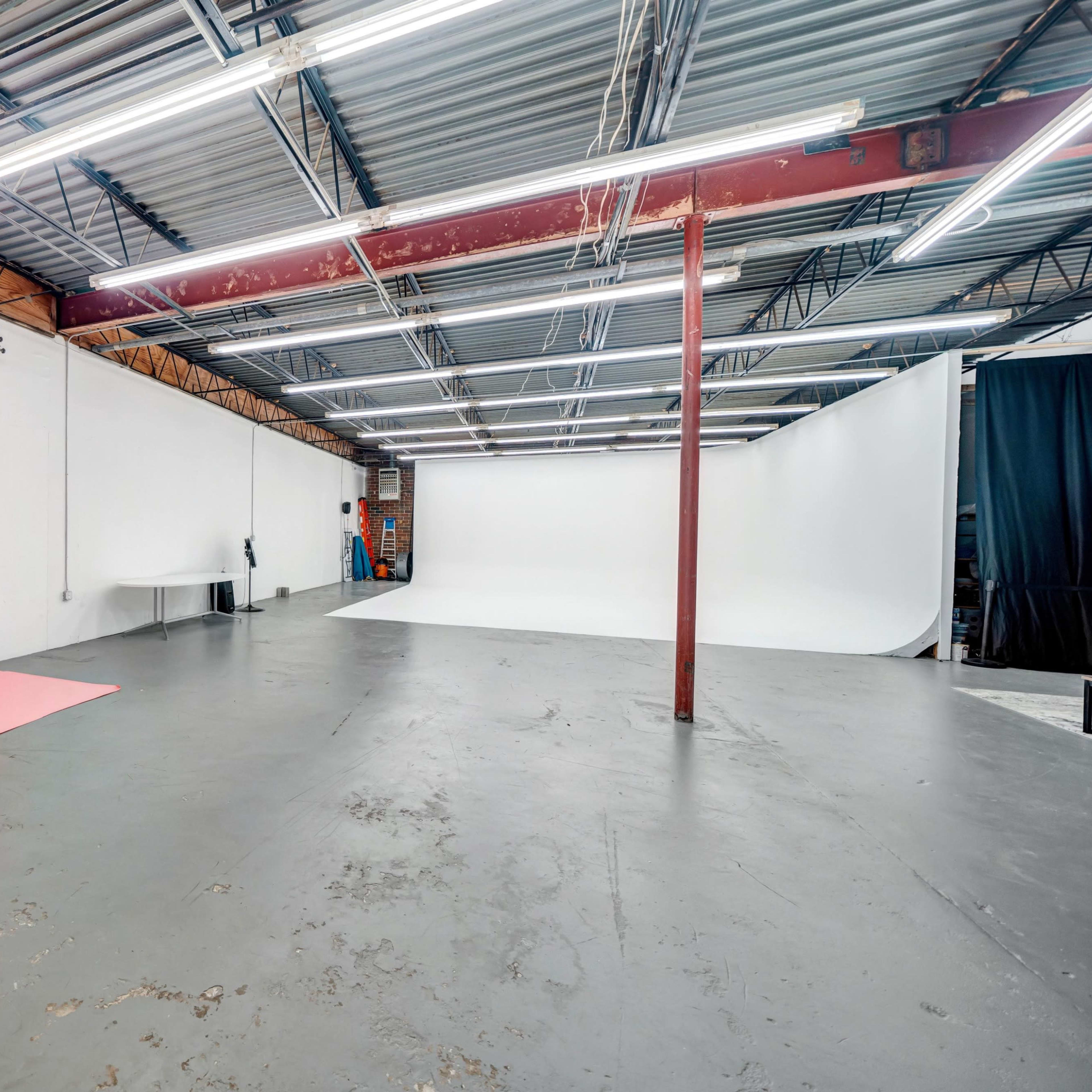 A spacious photography studio featuring two backdrop areas, one with a pink backdrop and another with a white backdrop, along with overhead fluorescent lighting.