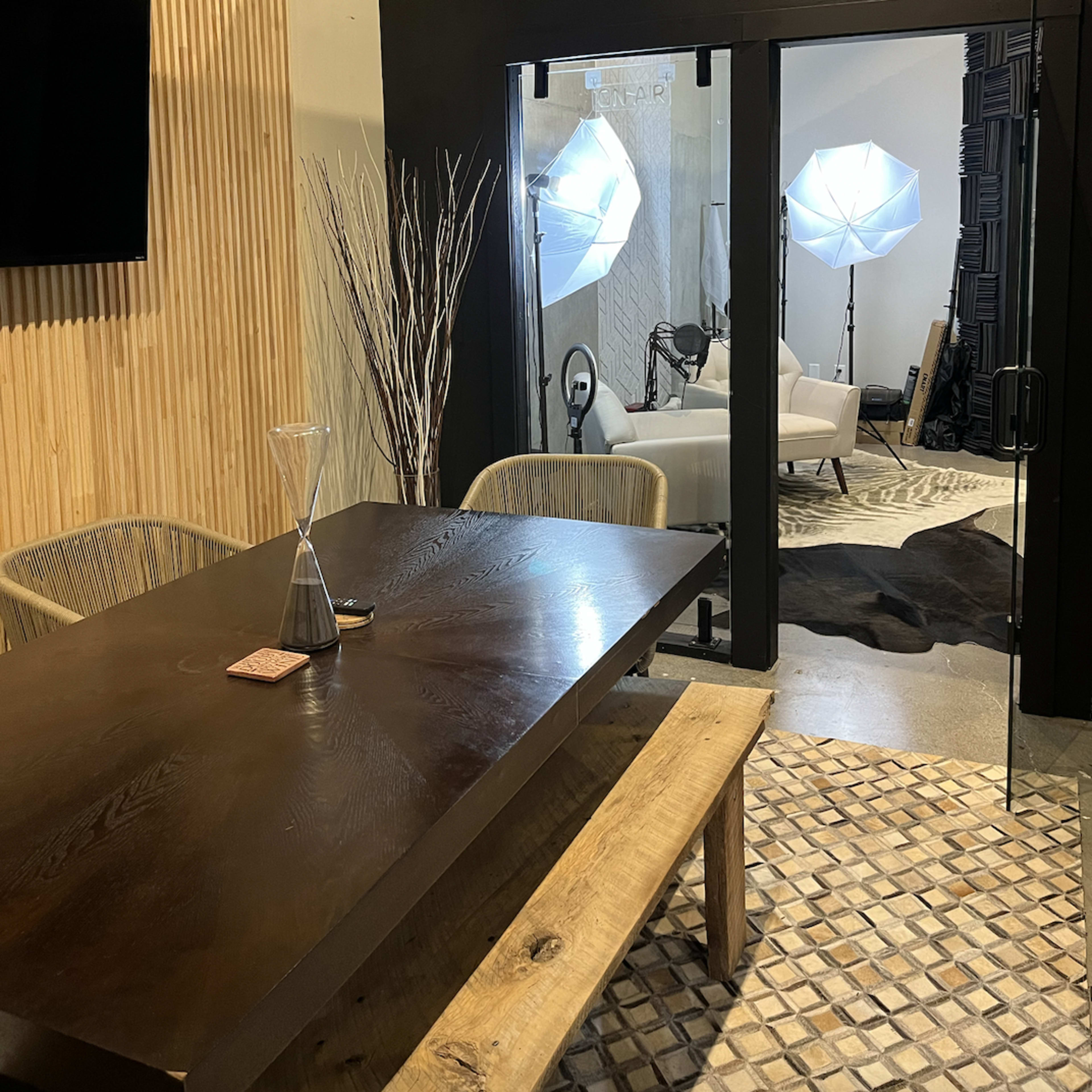 A modern meeting room with a large dark table, wooden accents, and bright studio lights illuminating a nearby seating area.