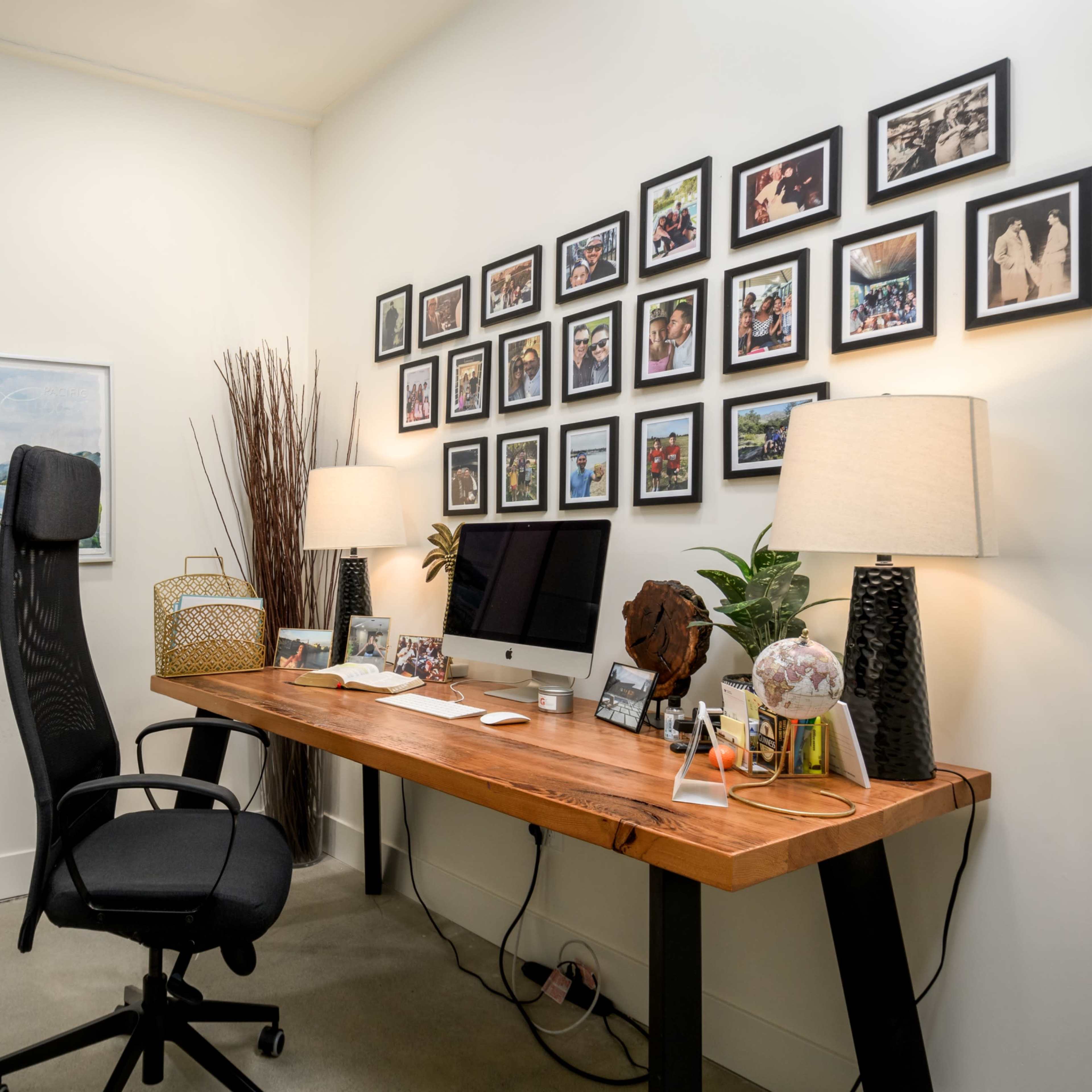 A modern office features a wooden desk with a computer, surrounded by framed photos on the wall, a large mirror, and decorative lamps.