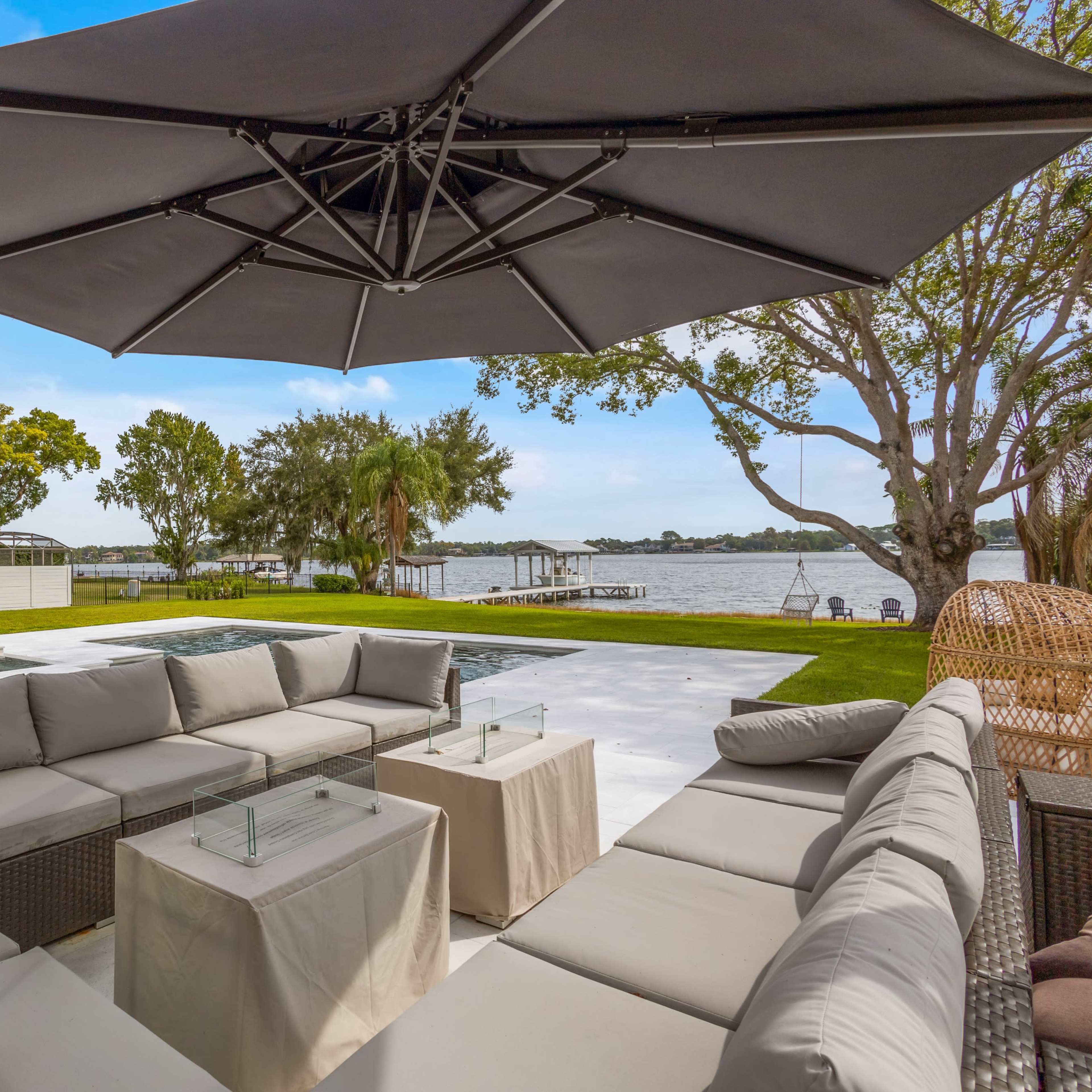 A modern outdoor seating area with a large umbrella overlooks a calm body of water surrounded by trees.