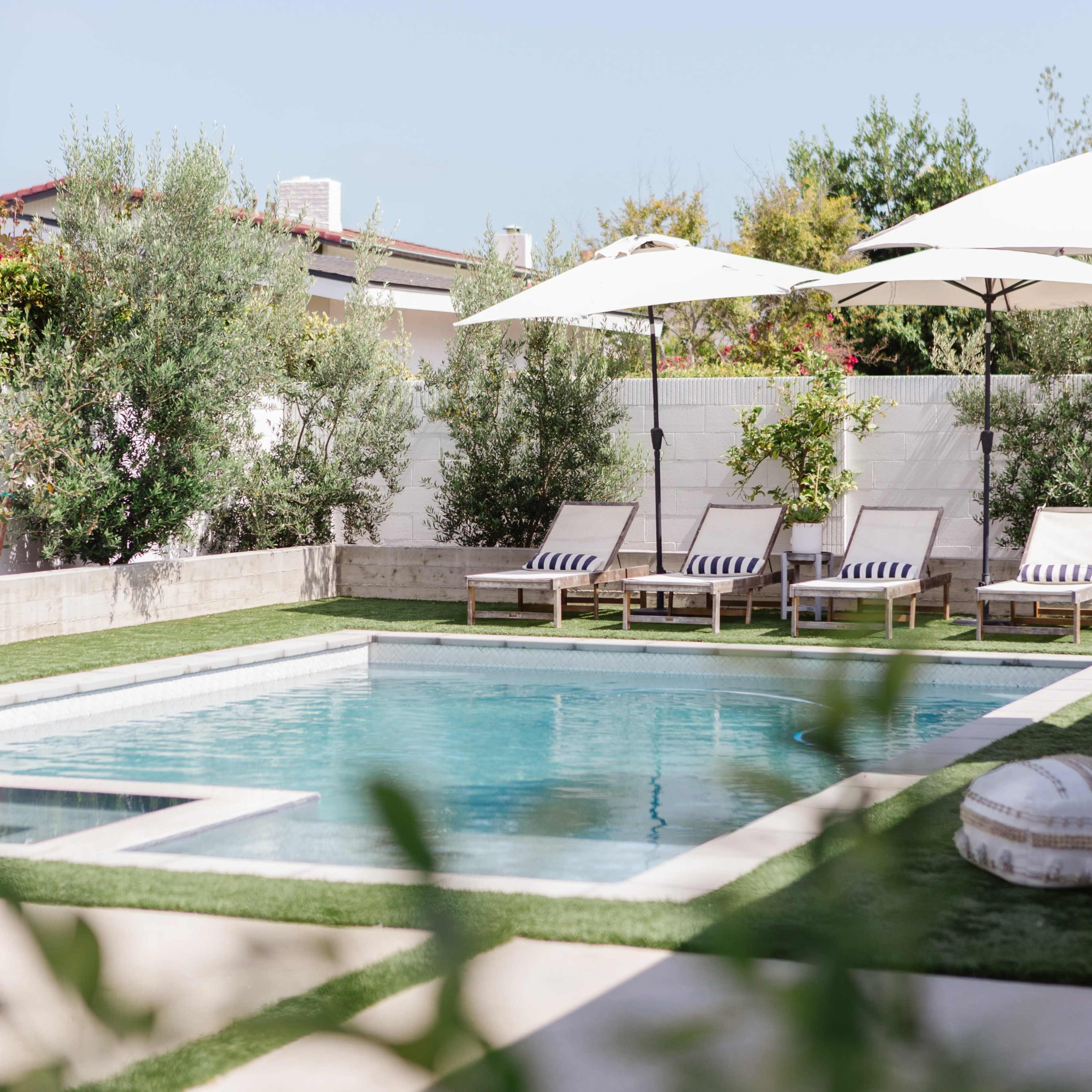 A rectangular swimming pool is surrounded by lounge chairs and umbrellas in a landscaped backyard.