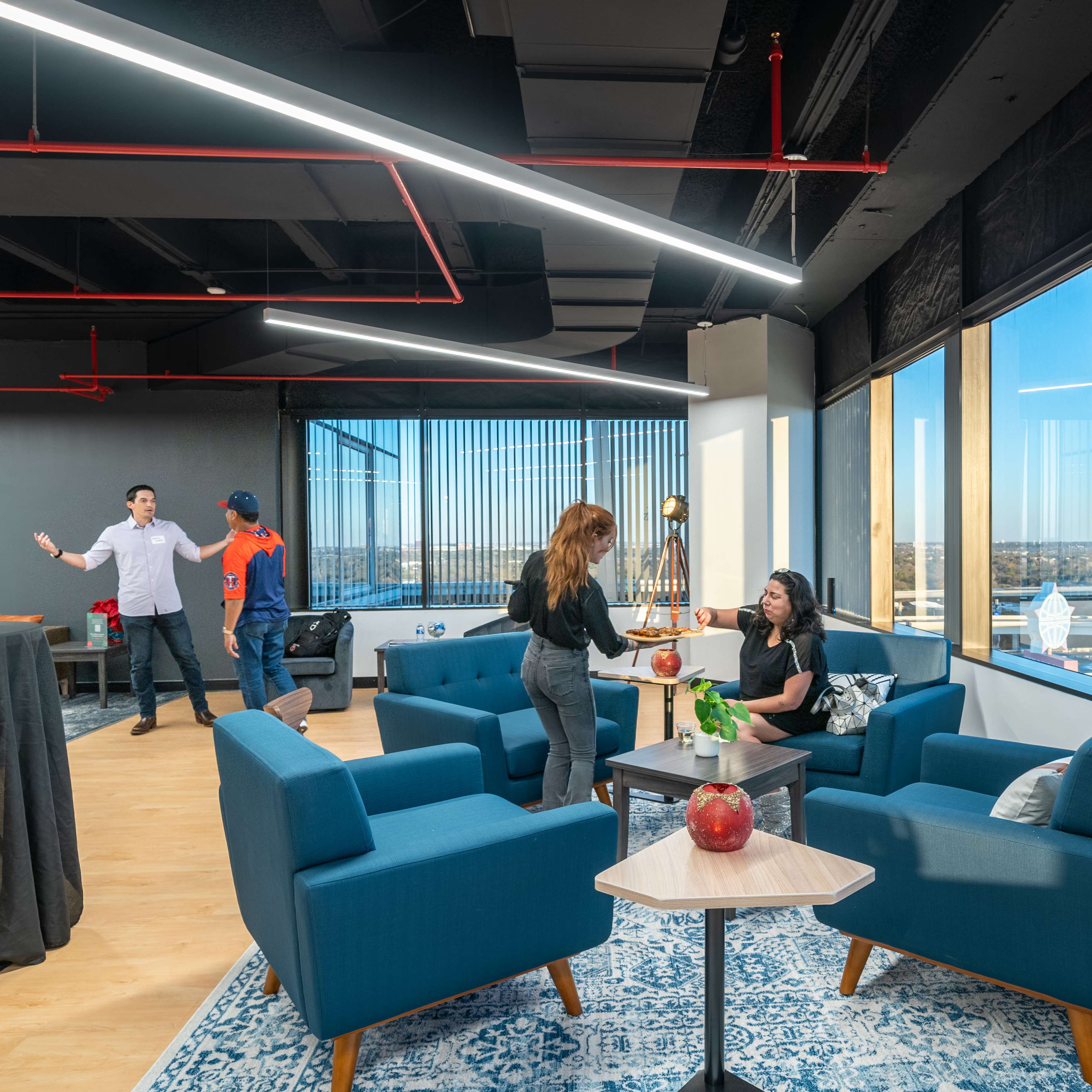 A modern office lounge features two people chatting at a table while a third person prepares a drink, with large windows showcasing a city view.