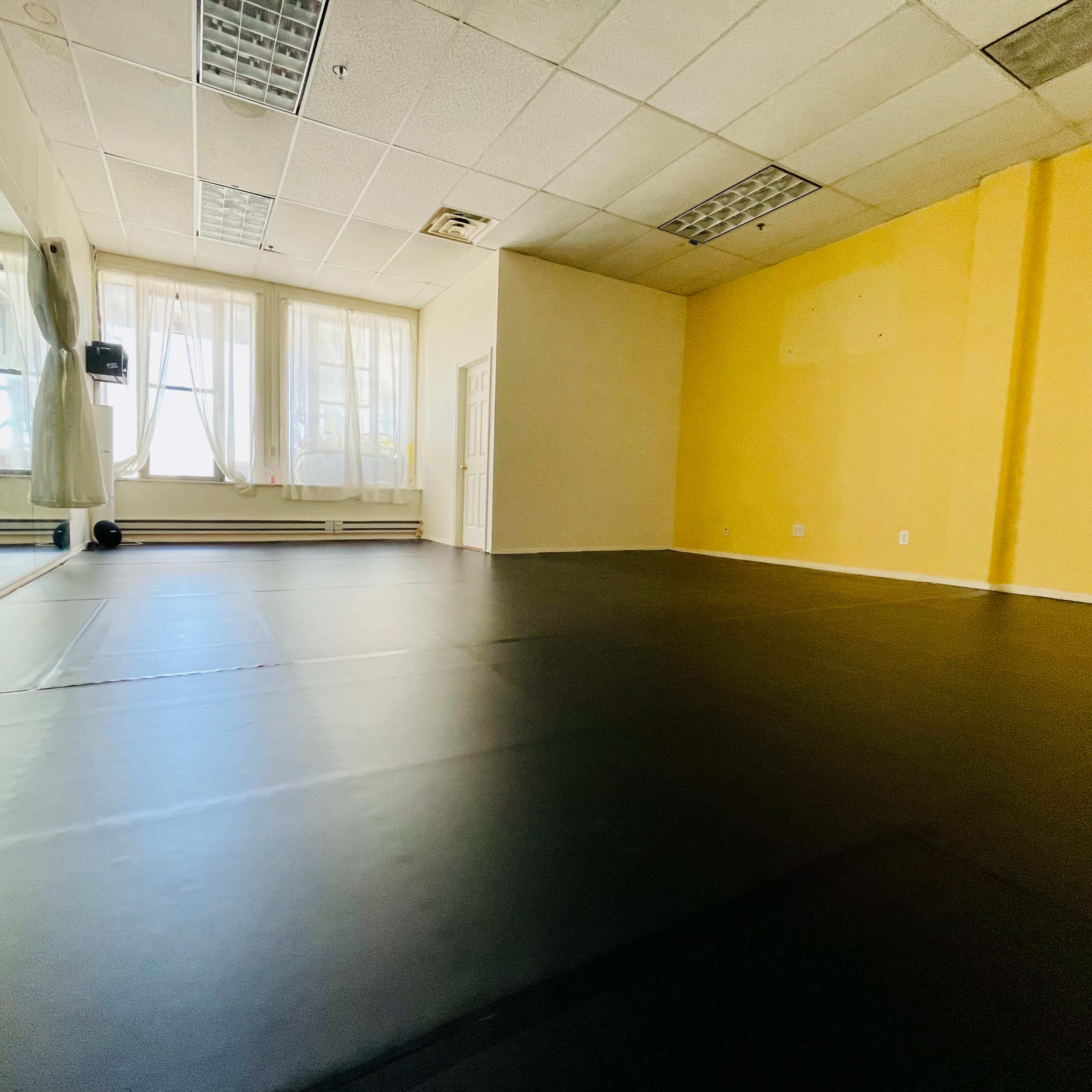 An empty room with black flooring, large windows, and a mirror along one wall.
