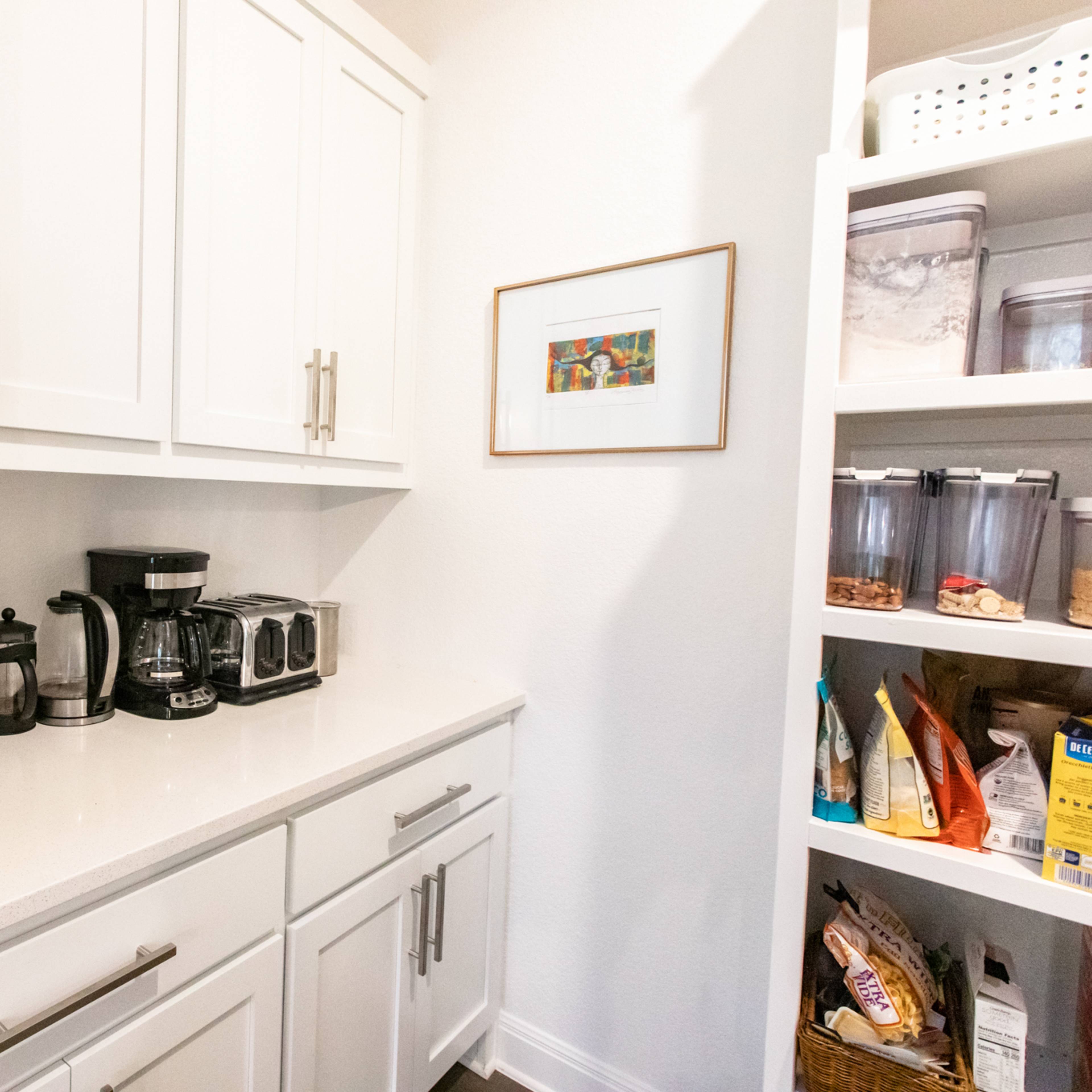 A kitchen pantry features white cabinetry, a countertop with coffee-making appliances, and organized shelves filled with food items and containers.