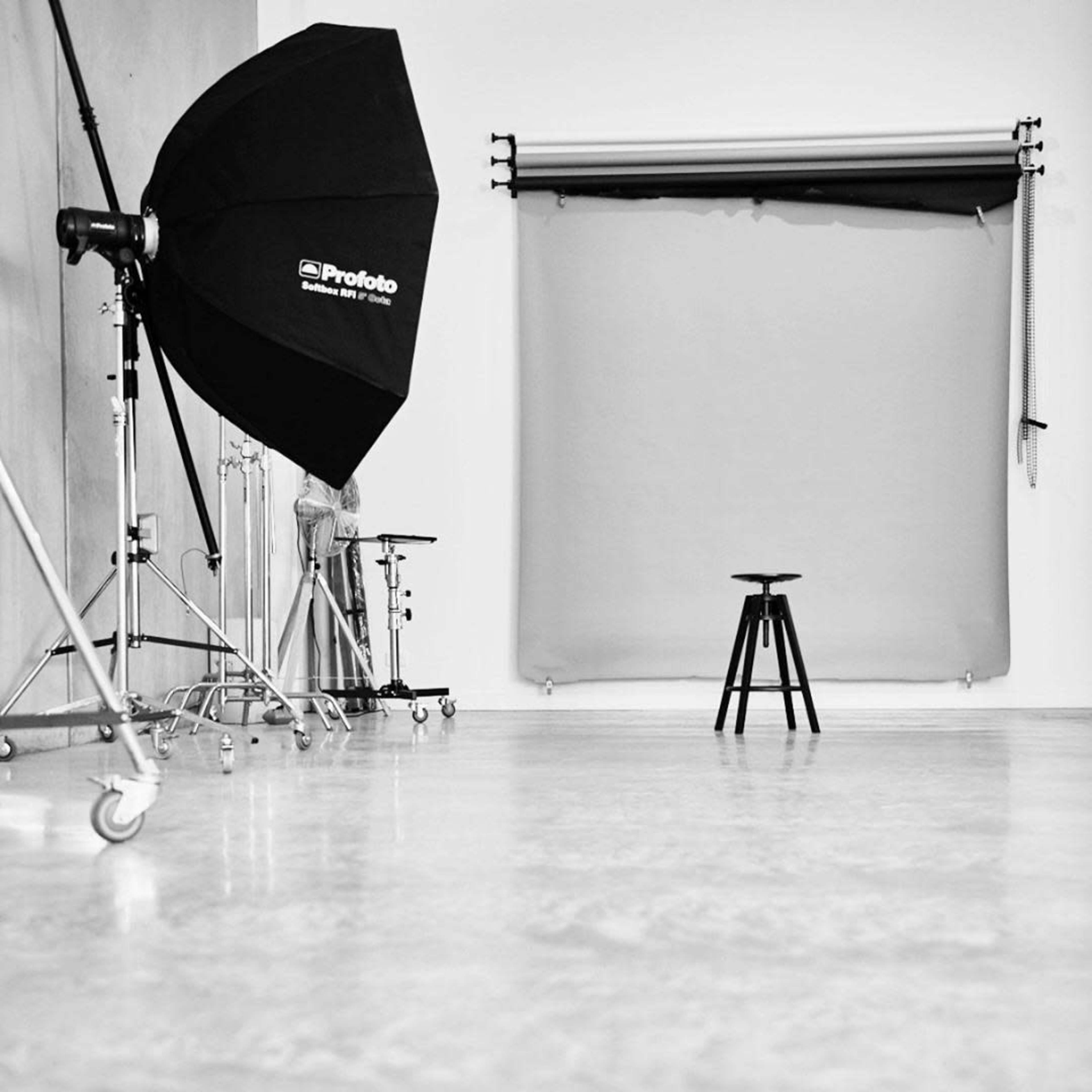 A photography studio with a black umbrella light, a gray backdrop, and a black stool in the center.