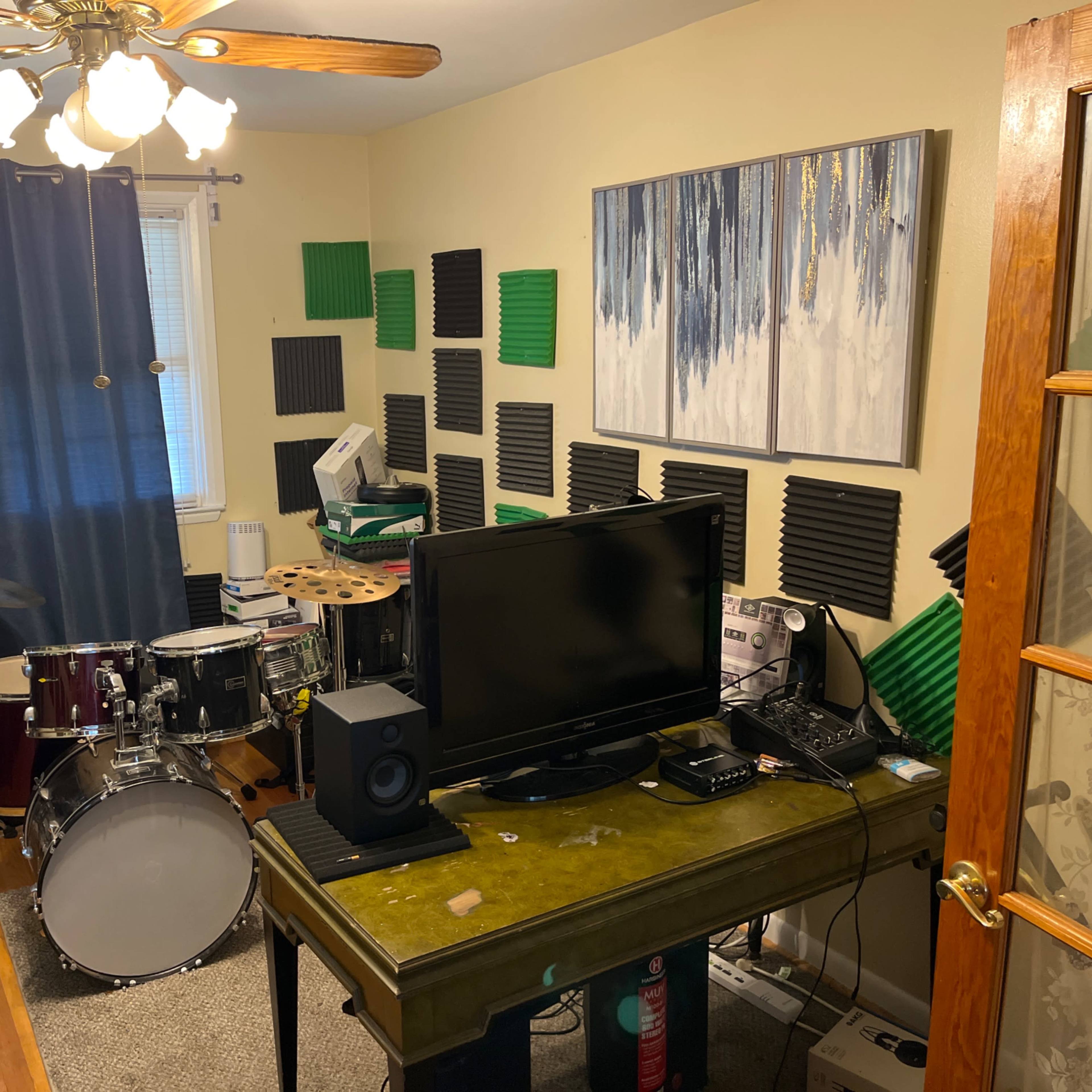 A small room features a drum set, a television, and a desk with electronic equipment, surrounded by soundproofing panels on the walls.