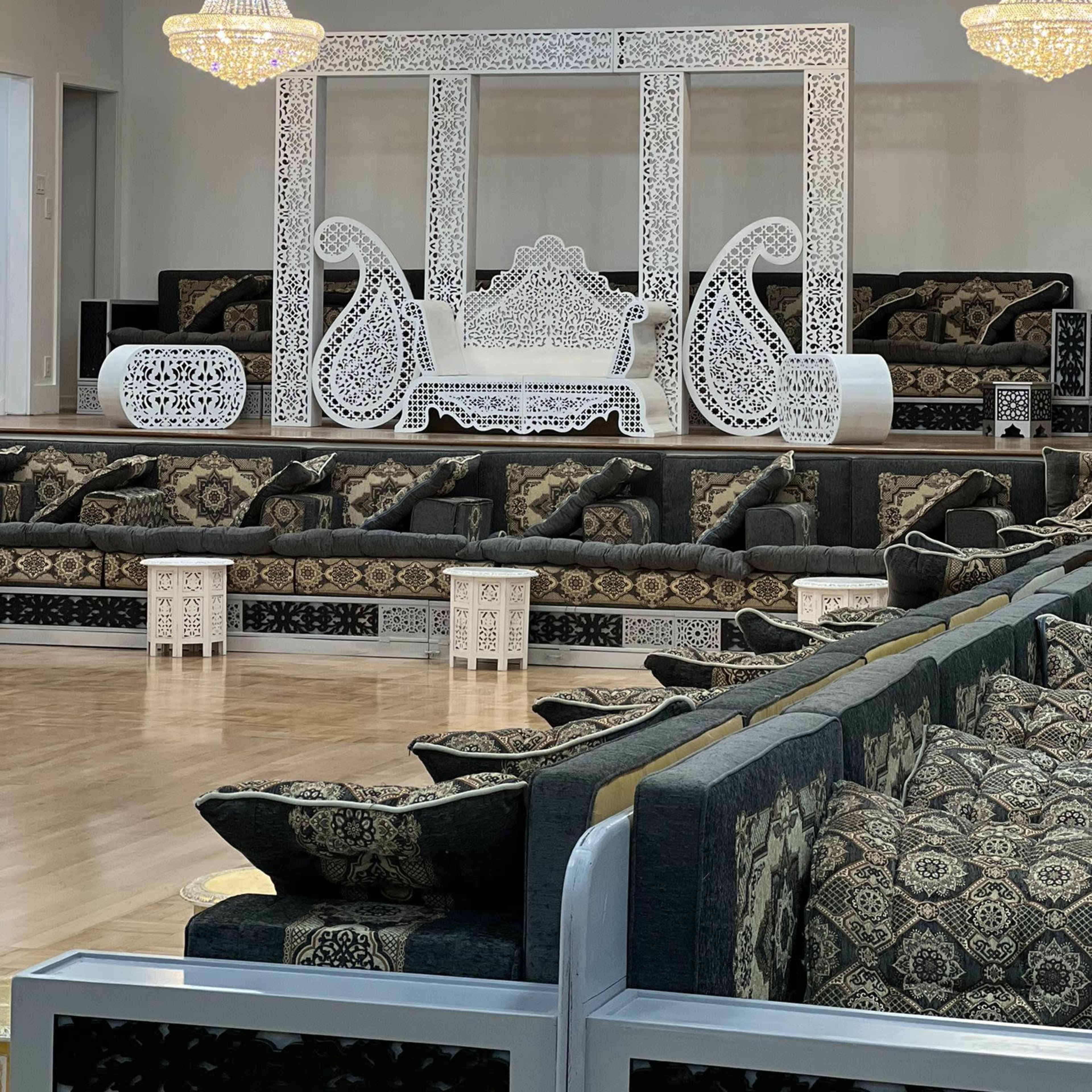 A spacious, elegantly arranged seating area featuring intricately designed white furniture and plush patterned cushions under two hanging chandeliers.