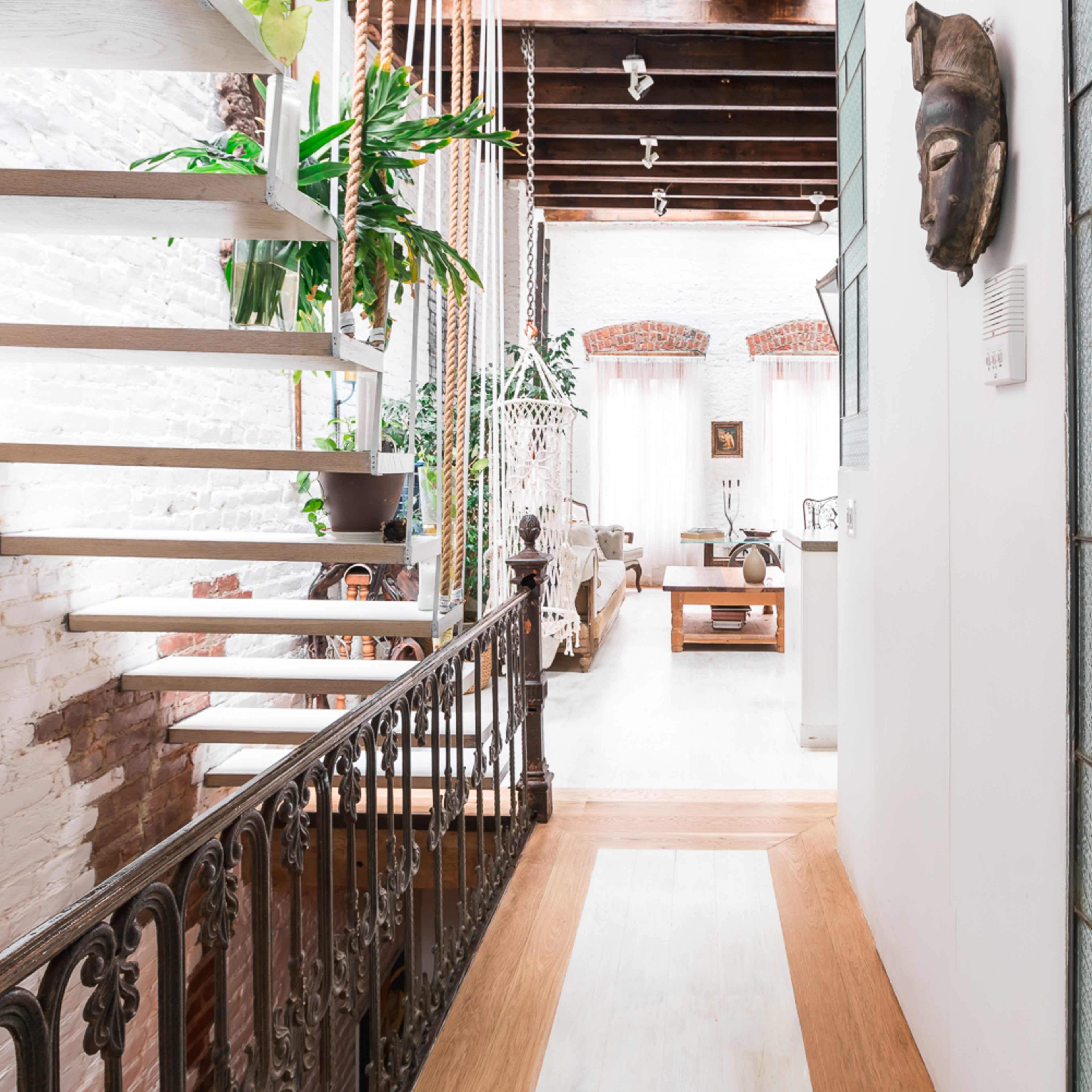 A narrow corridor features a staircase leading to an upstairs area, flanked by plants and open spaces with light streaming in through large windows.