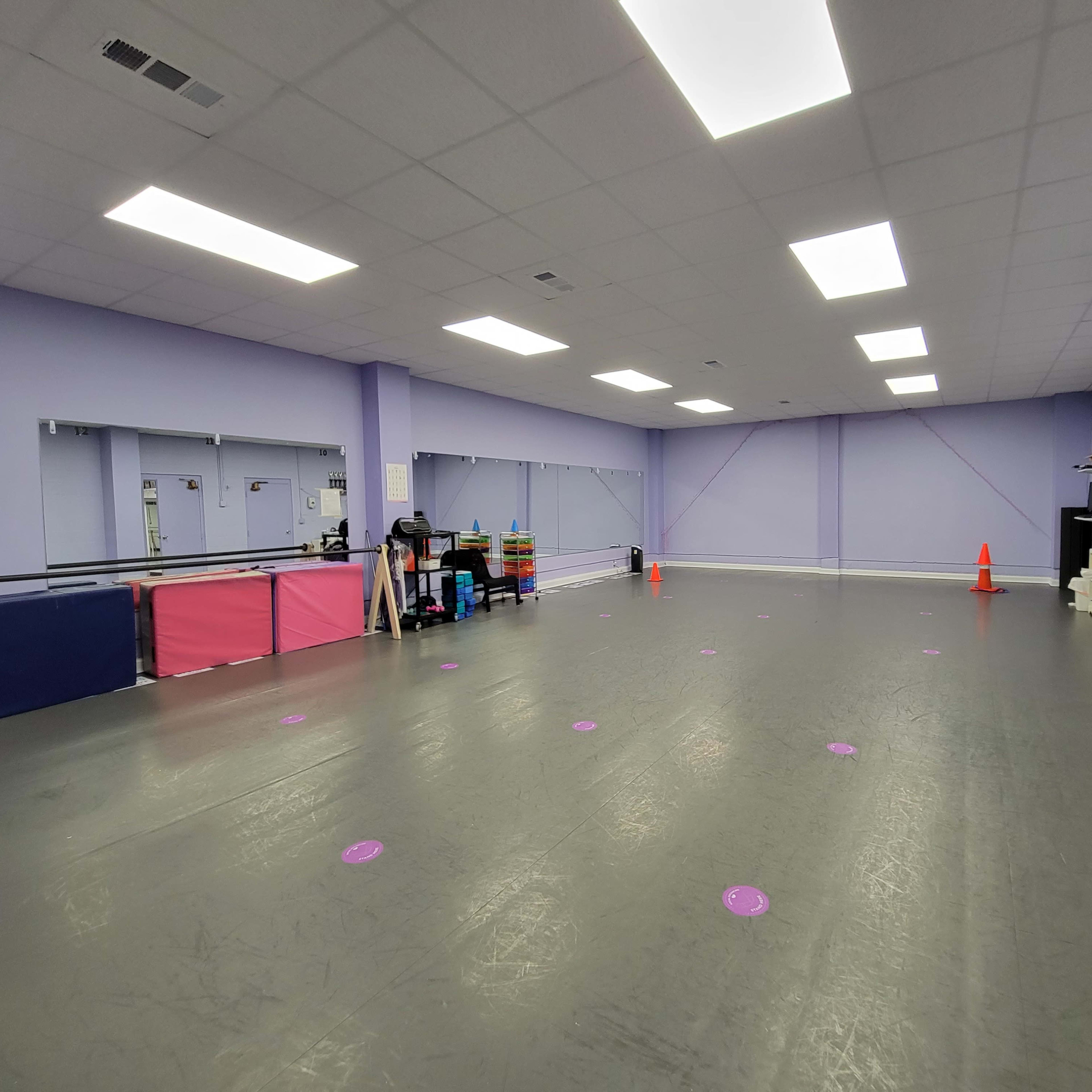 A spacious dance studio with a gray floor, purple walls, and mirrors, featuring some storage equipment and pink mats along one side.