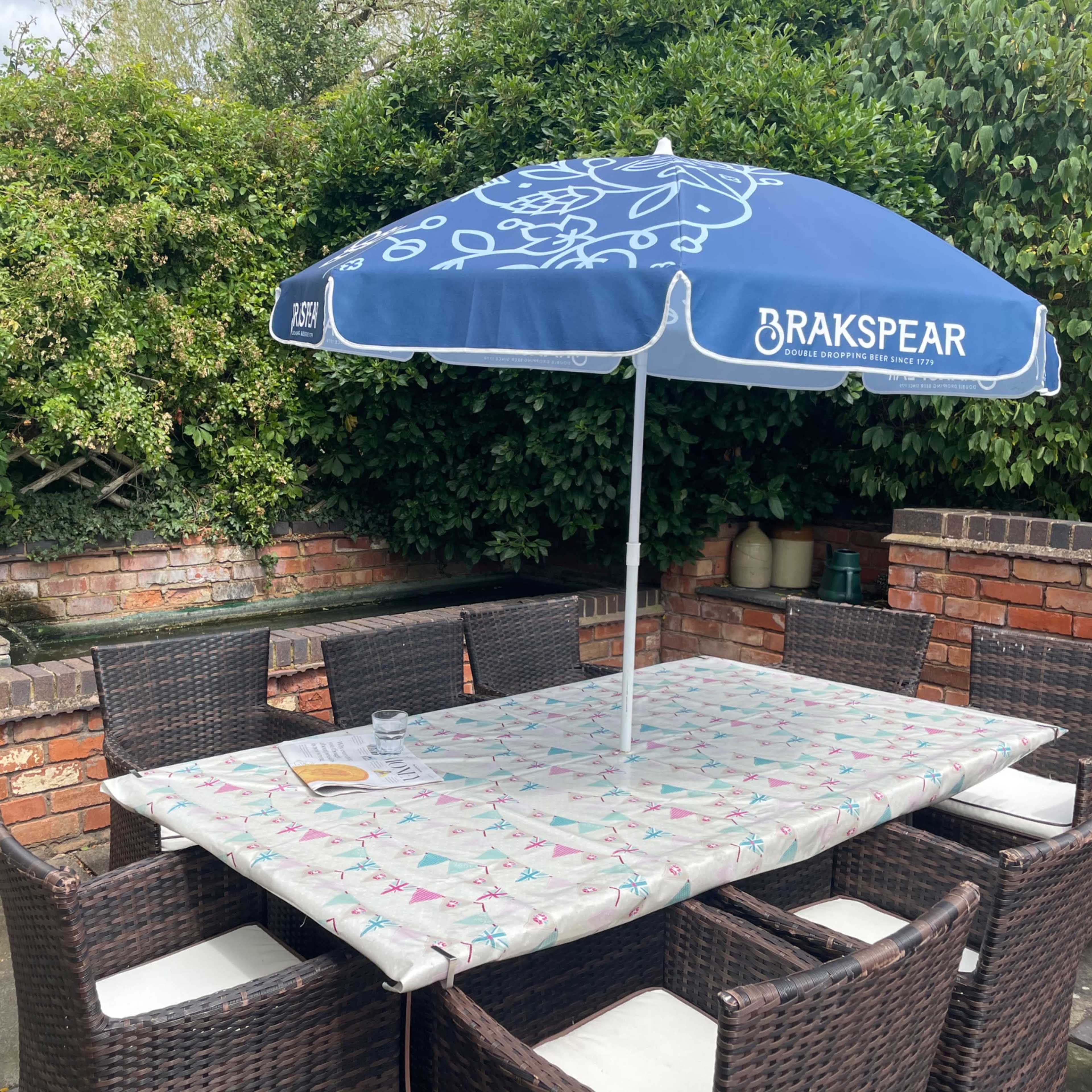 A patio table with a patterned tablecloth is surrounded by eight wicker chairs under a blue umbrella next to a greenery-covered wall and a water feature.